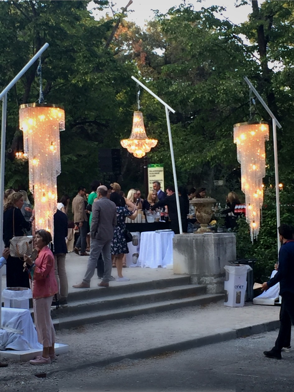 Un événement en plein air avec des gens socialisant, décorations lumineuses, lustre suspendu et stands avec serviettes. Atmosphère élégante et festive.