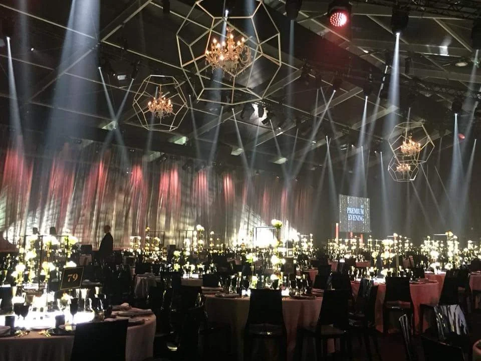 Salle de banquet élégante avec des tables rondes, ornées de vaisselle, couverts et fleurs, éclairée par des chandeliers et des lumières de scène.