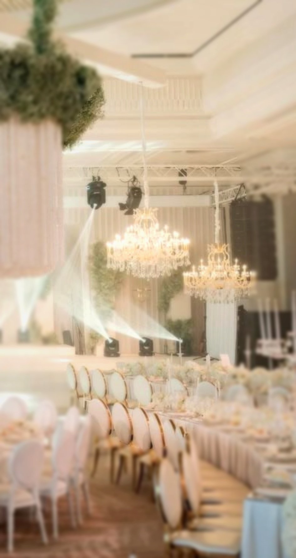 Salle de réception élégante avec chandeliers, tables et chaises, éclairage de scène, et décoration florale, prête pour un événement ou un banquet.