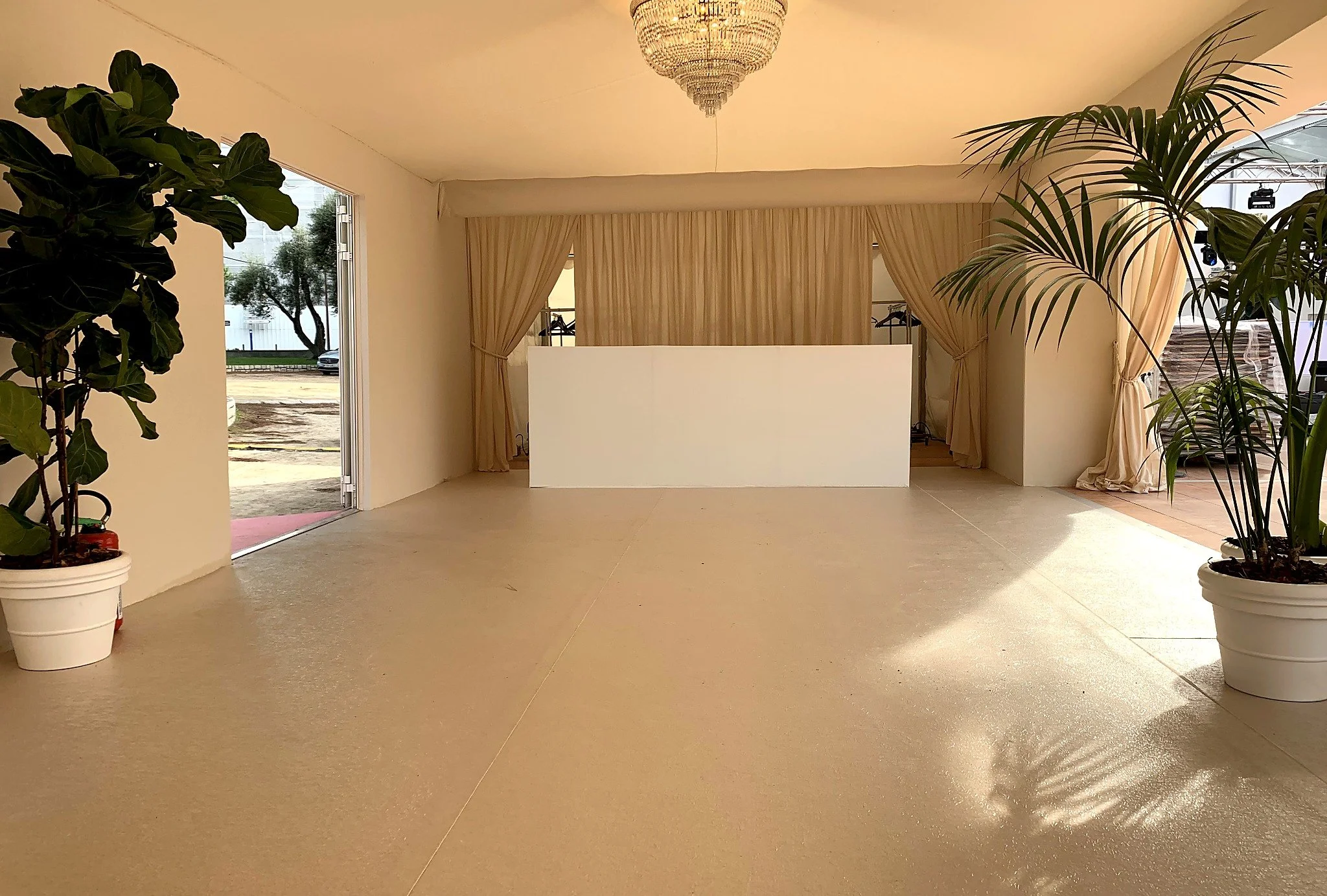 Intérieur d'une salle avec des plantes en pot, un lustre suspendu au plafond, et des rideaux beiges devant une ouverture ou un espace de projection. La lumière naturelle entre par une porte ou fenêtre ouverte, montrant une rue avec des arbres et une 