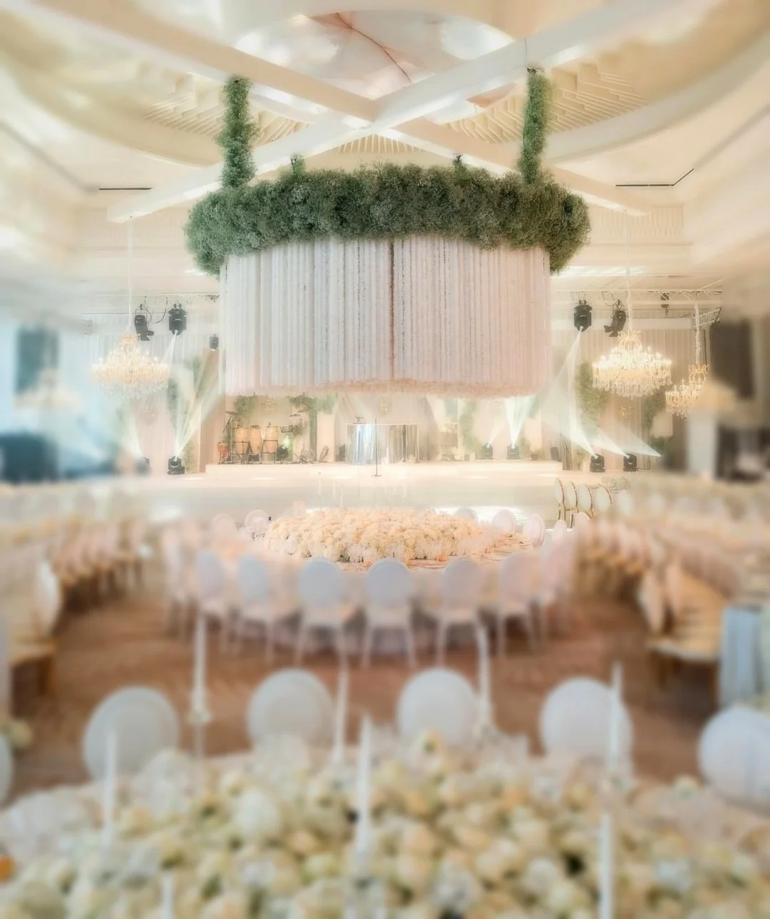 Salle de réception élégante avec un plafond orné de chandeliers, un centre de table suspendu en forme de grand cylindre couvert de verdure, et des tables décorées de fleurs pour un événement spécial.