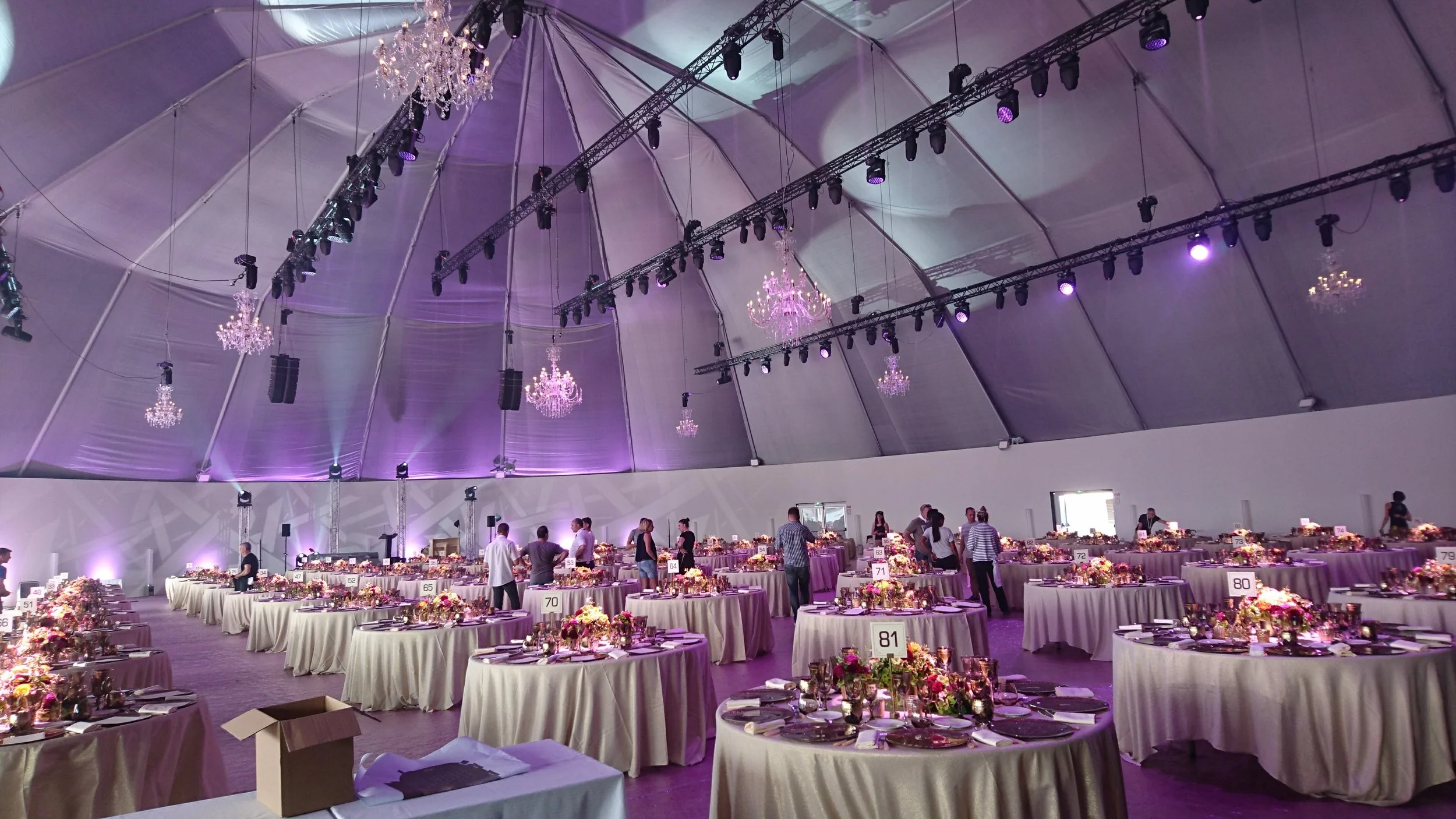 Grande salle préparée pour un événement ou un banquet avec de nombreuses tables rondes décorées de nappes blanches et de compositions florales, sous un plafond haut avec des chandeliers et un éclairage tamisé violet.
