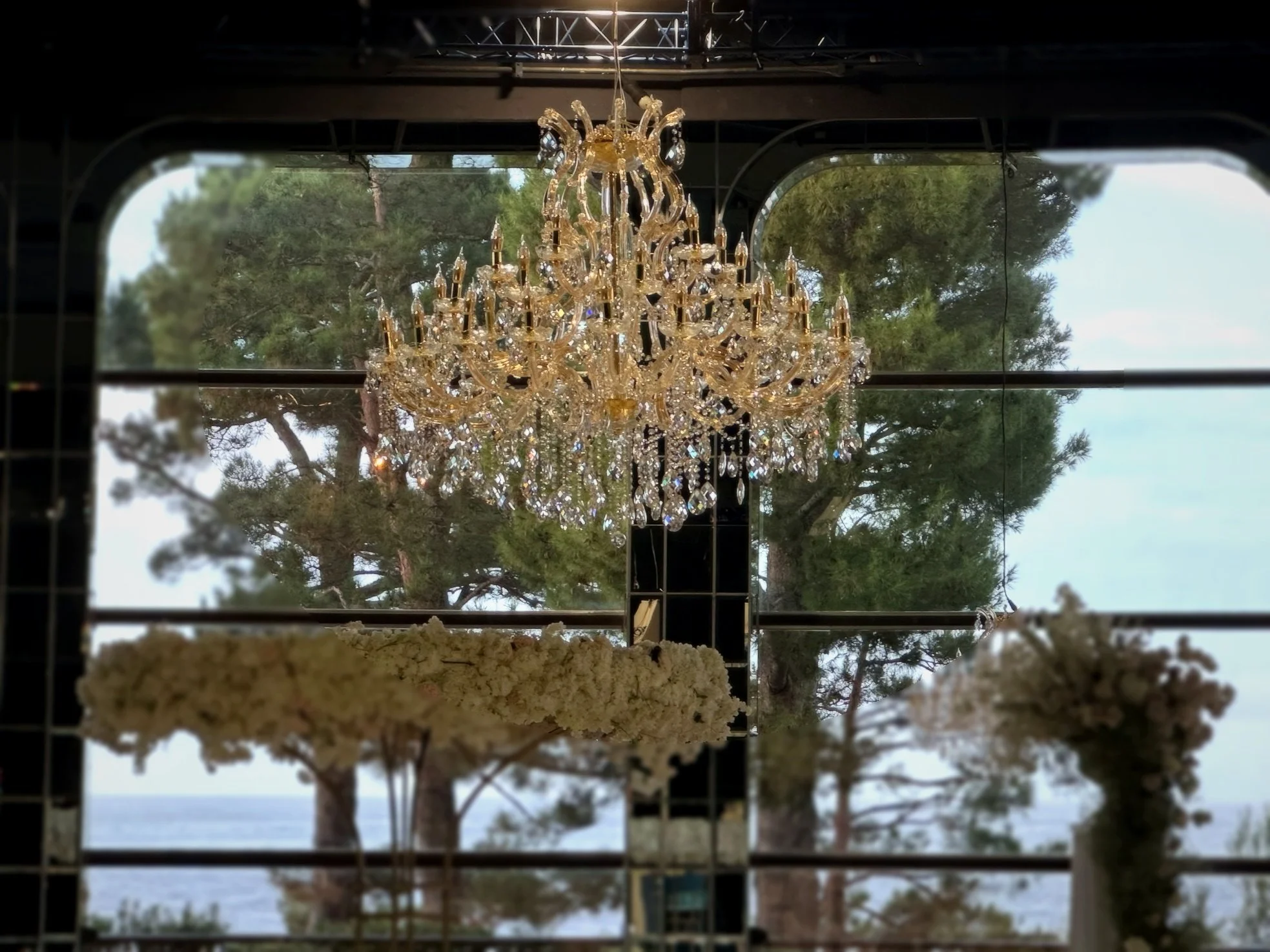 Un lustre en cristal suspendu avec des grandes fenêtres derrière, offrant une vue sur des arbres et la mer.
