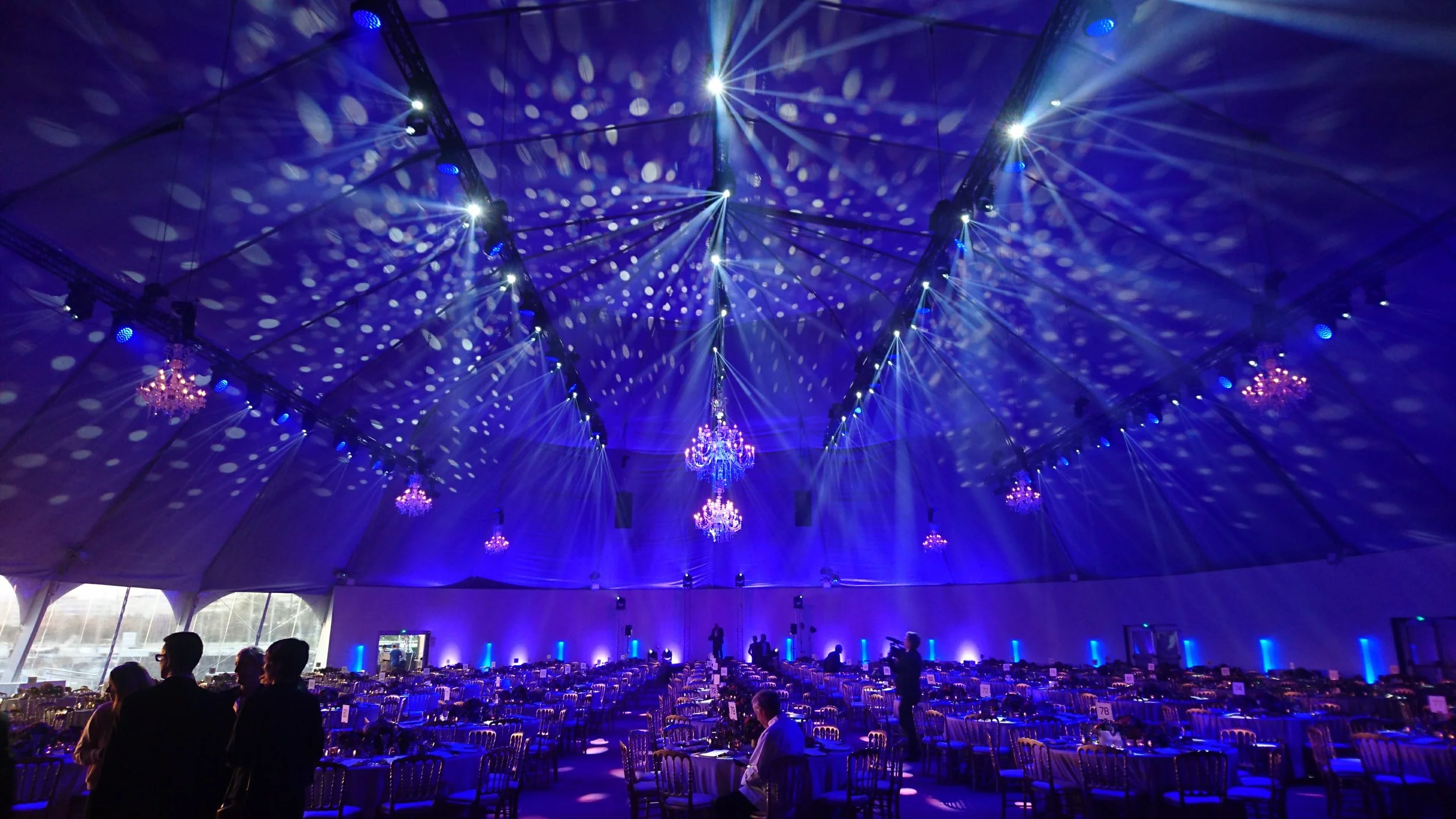 Intérieur d'une grande tente décorée avec des éclairages bleus, chandeliers suspendus, tables et chaises pour un événement ou une réception, ambiance festives
