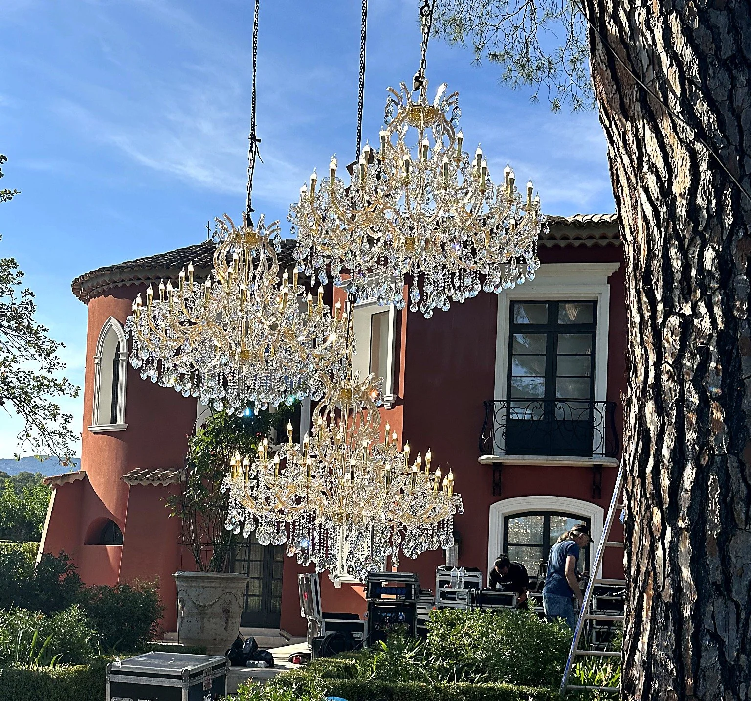 Deux lustres en cristal suspendus en dehors d'une maison, avec quelques personnes et du matériel d'installation au sol.