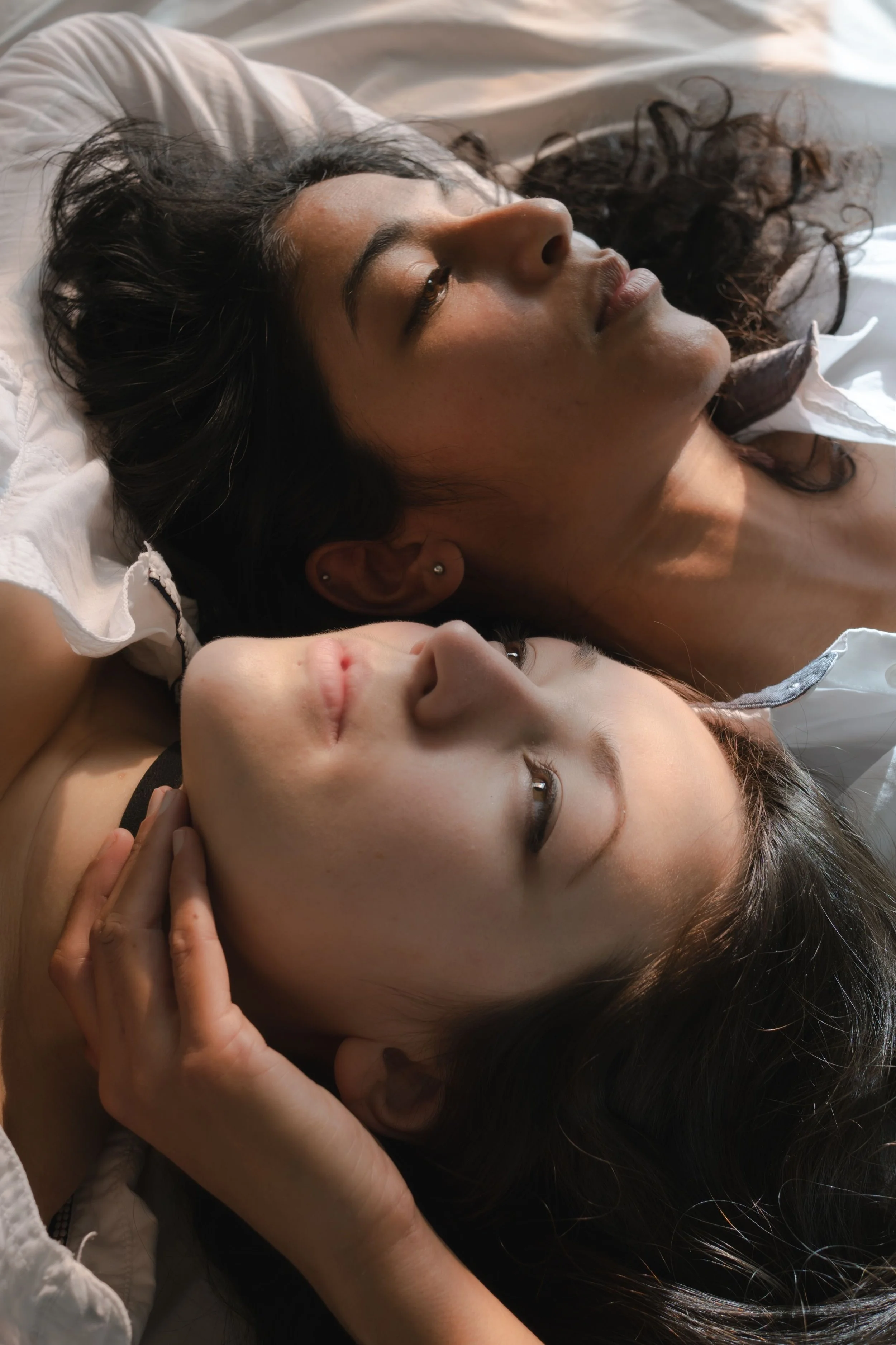 Two women lying close together, one with long dark hair and the other with light brown hair, are posing on a bed with sunlight illuminating their faces.