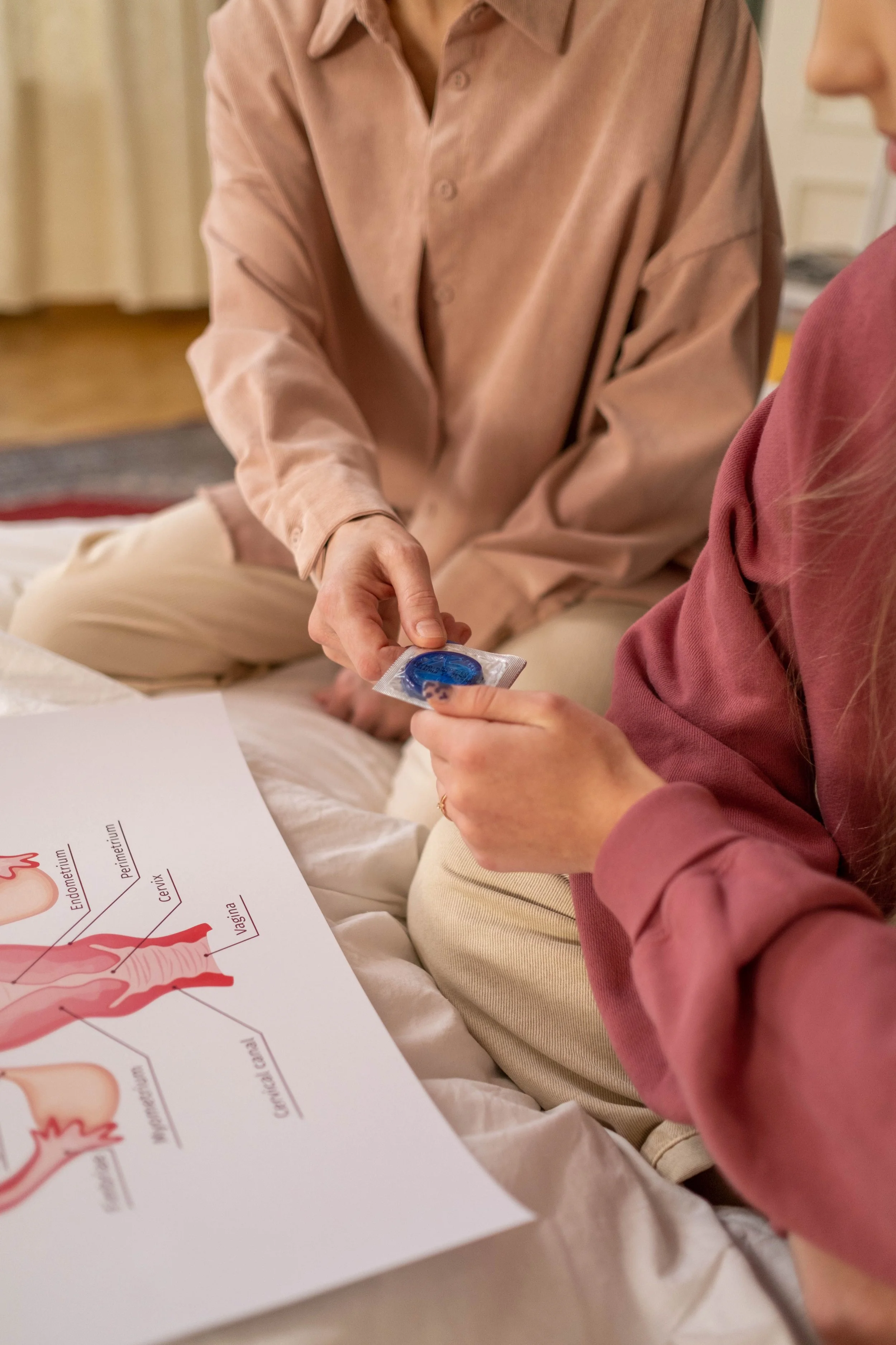 Two people sitting on a bed, one person handing a condom to the other, with an anatomical diagram of the female reproductive system on the surface.