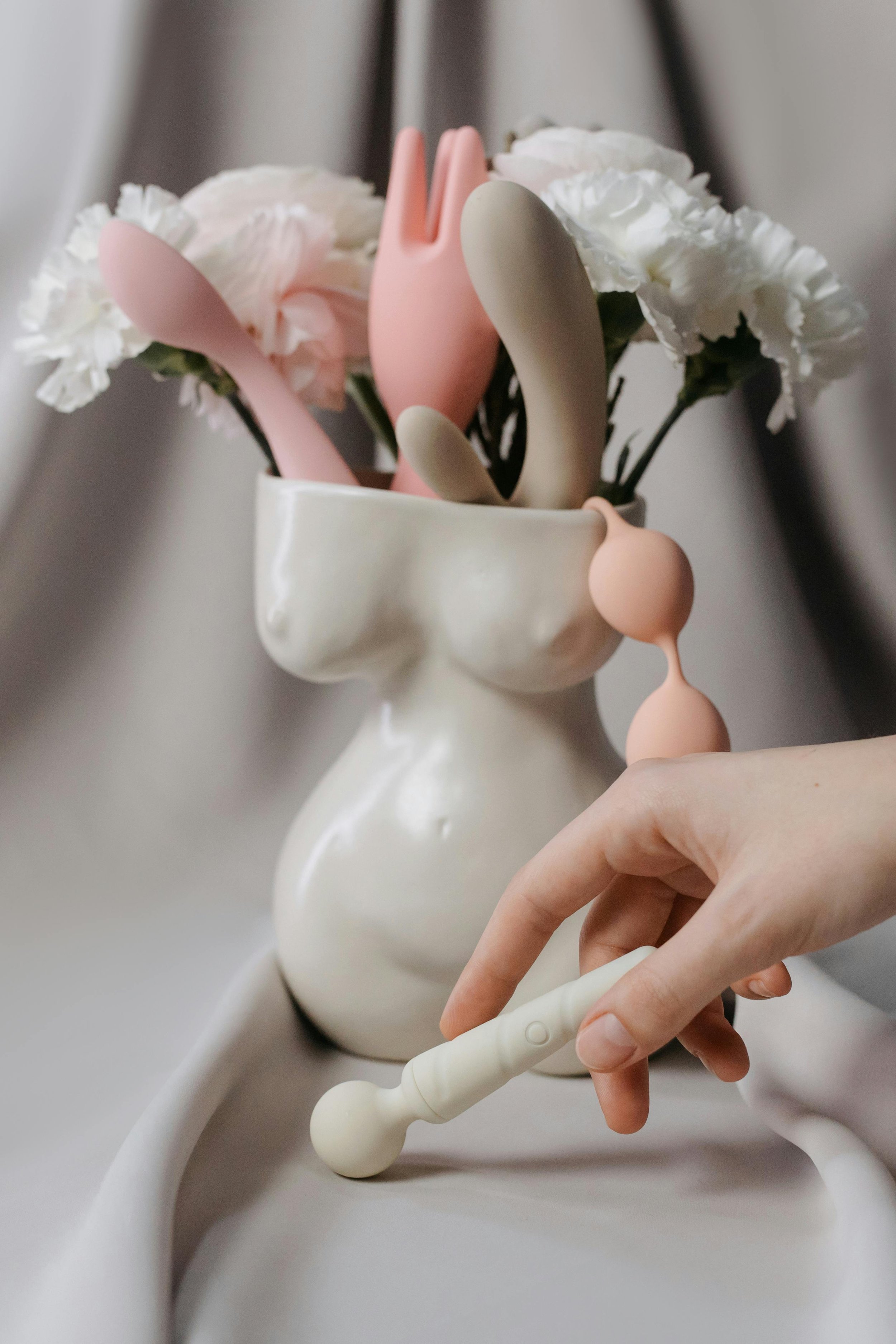 A white vase with artificial white flowers and pastel-colored silicone baby pacifiers inserted among the flowers. A person's hand is reaching to adjust or remove a pacifier.