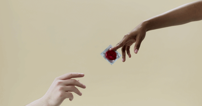 Two hands, one with pale skin and one with darker skin, engage in an exchange. The darker-skinned hand is dropping a condom with a red heart shape inside onto the palm of the pale-skinned hand, against a plain beige background.