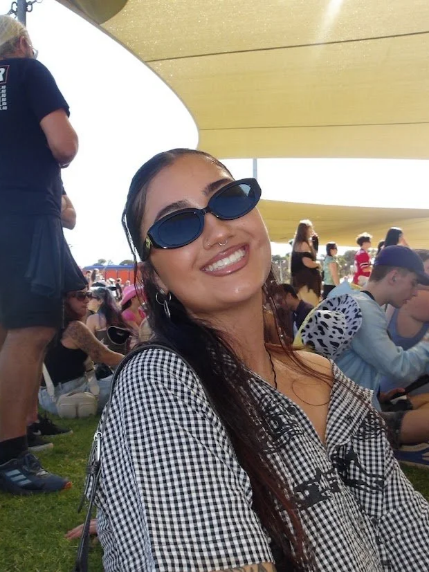Smiling woman wearing black sunglasses and a black-and-white checkered shirt, sitting outdoors at a crowded event under a large yellow canopy
