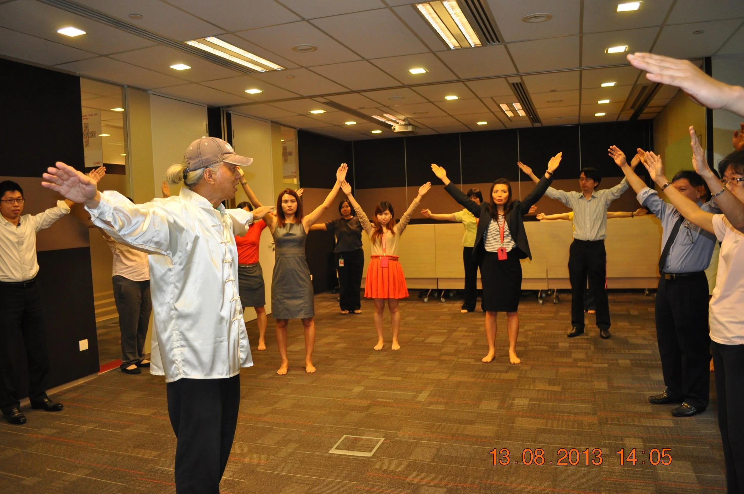 A group of people in a large room participating in a group activity, standing in a circle with arms raised and hands open. The instructor, at the front, appears to be leading the session, demonstrating a pose with arms outstretched.