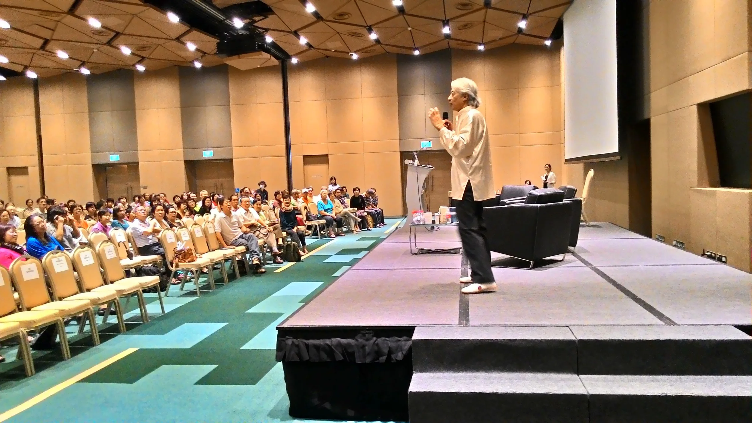 A speaker in traditional attire giving a talk on stage at a conference, with an audience seated and listening attentively in a large, well-lit auditorium.