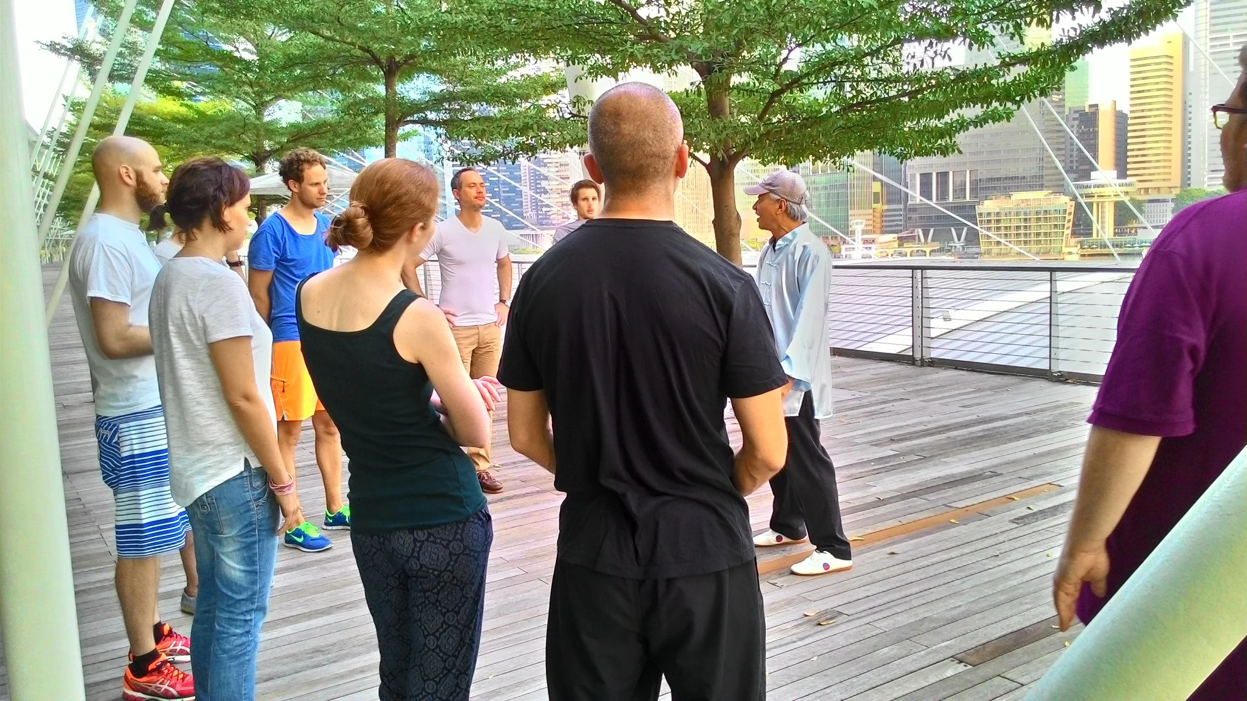 A group of people standing outdoors on a wooden deck, listening to an older man speaking. Trees and city buildings are visible in the background.