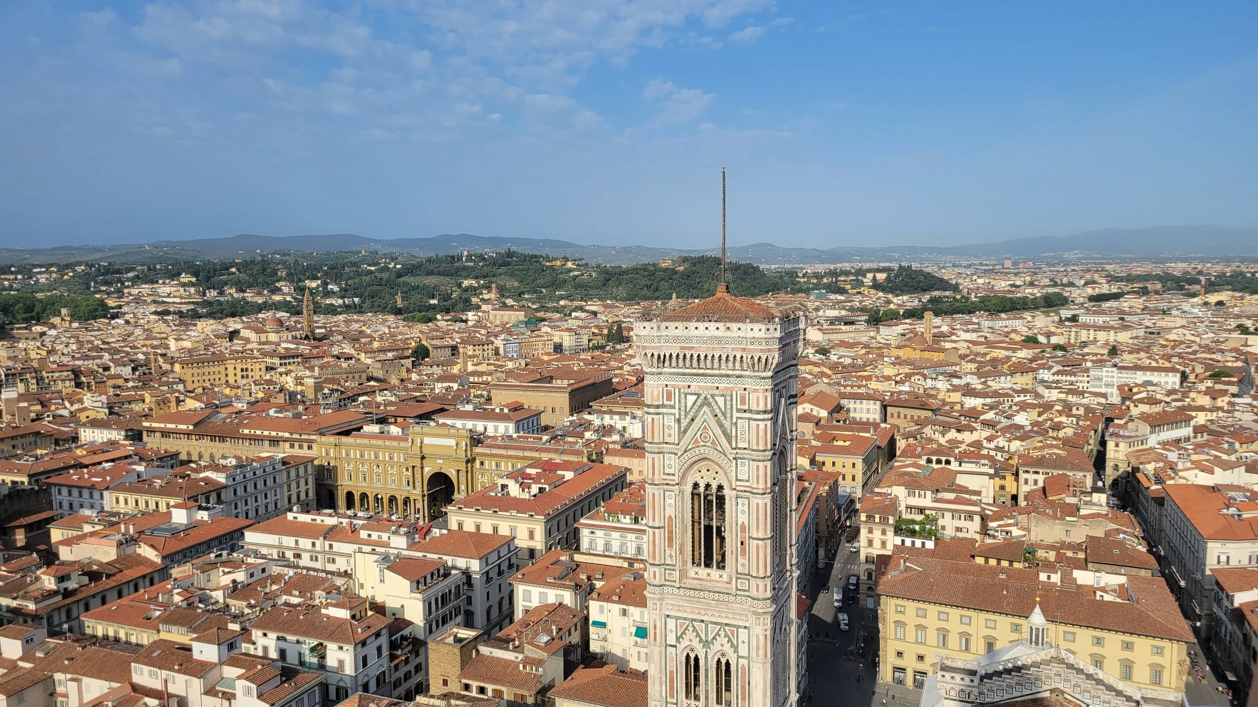 Aerial view of Florence, Italy, featuring the Florence Cathedral's bell tower with detailed marble design and the cityscape with terracotta rooftops and rolling hills in the background.