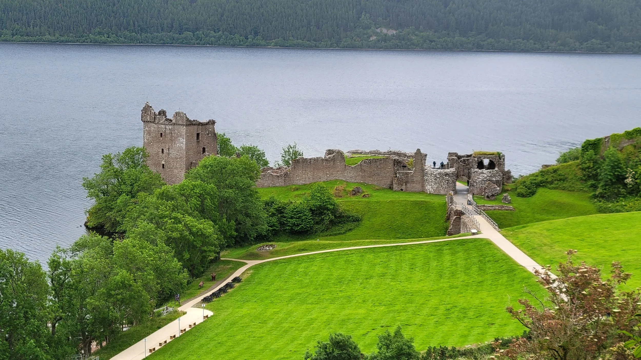 A historic stone castle located on a lush green landscape by a large body of water, with a pathway leading to the castle and surrounded by trees.