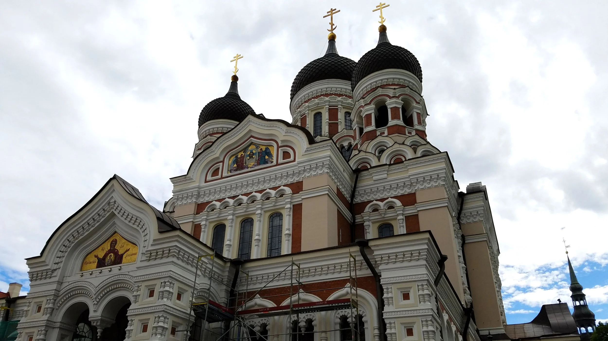 A large church with ornate architecture, three onion domes topped with gold crosses, and religious mosaics above the entrance, under an overcast sky. Tallin, Estonia