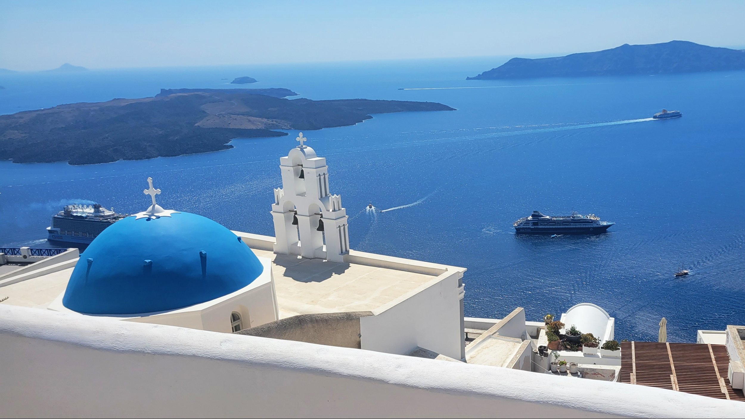 View of the Aegean Sea from Santorini, Greece, with blue-domed churches, white buildings, and boats on the water.