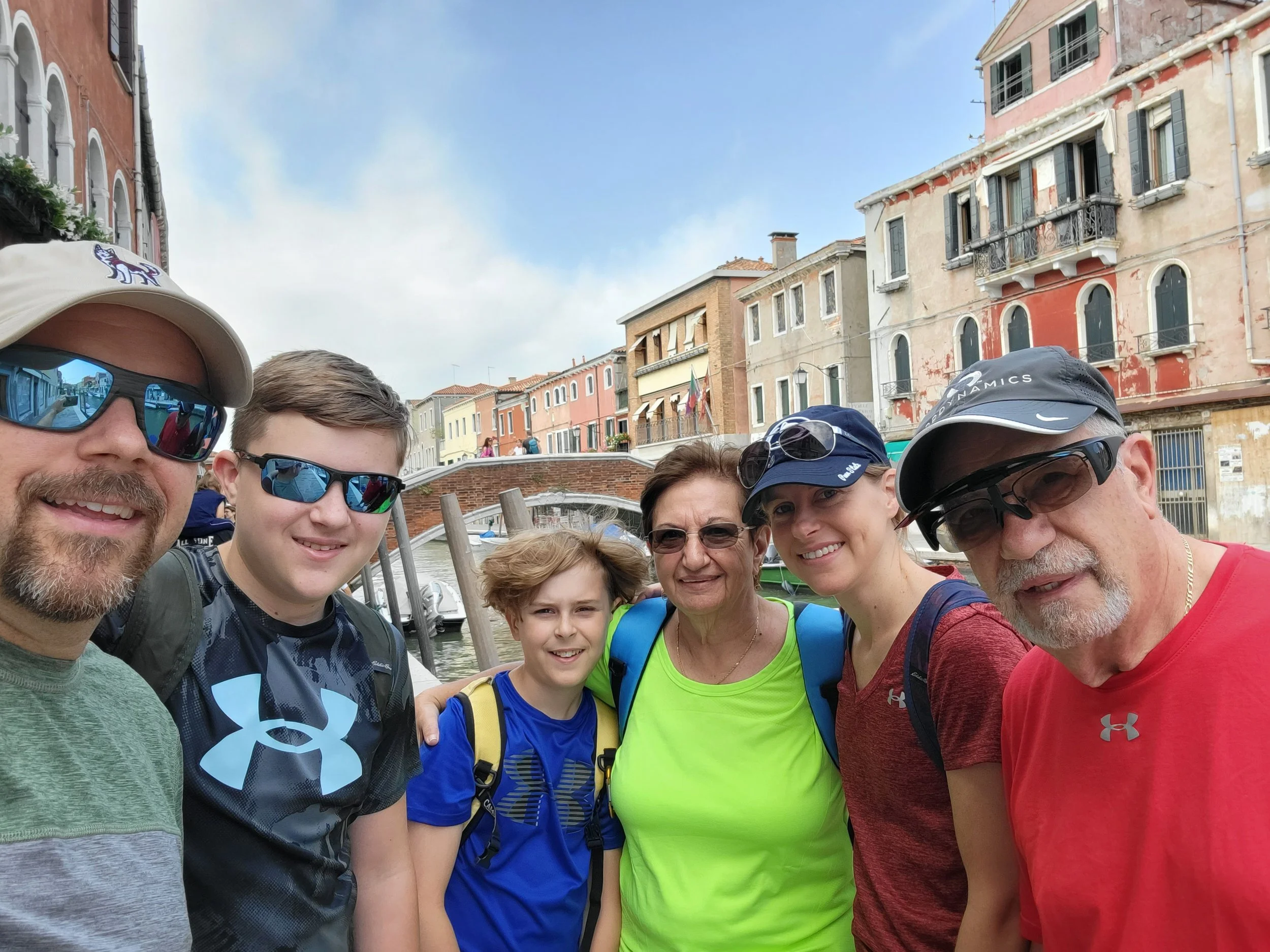 Six family members across three generations pose for a selfie in sun hats and sunglasses, with a canal and colorful older buildings behind them in Venice, Italy.
