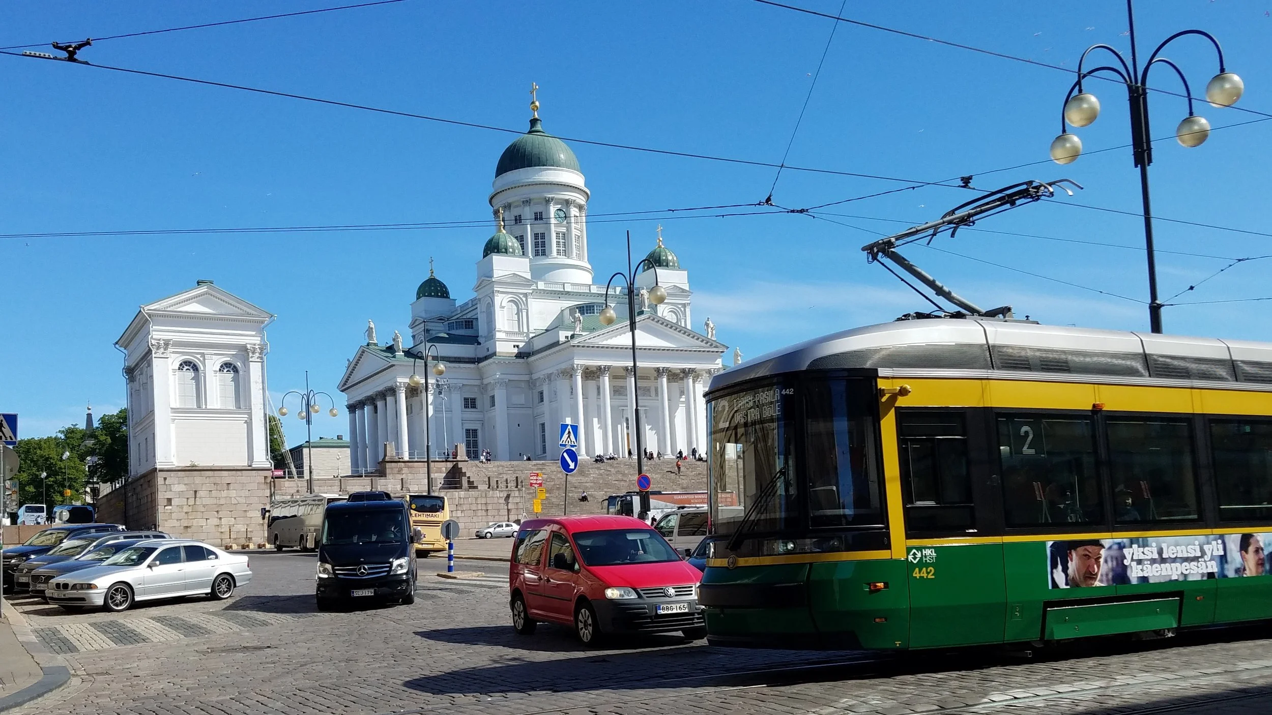 A city scene with a historic white church with green domes in the background, colorful cars parked, a trolleybus in the foreground, and a clear blue sky overhead. Helsinki, Finland