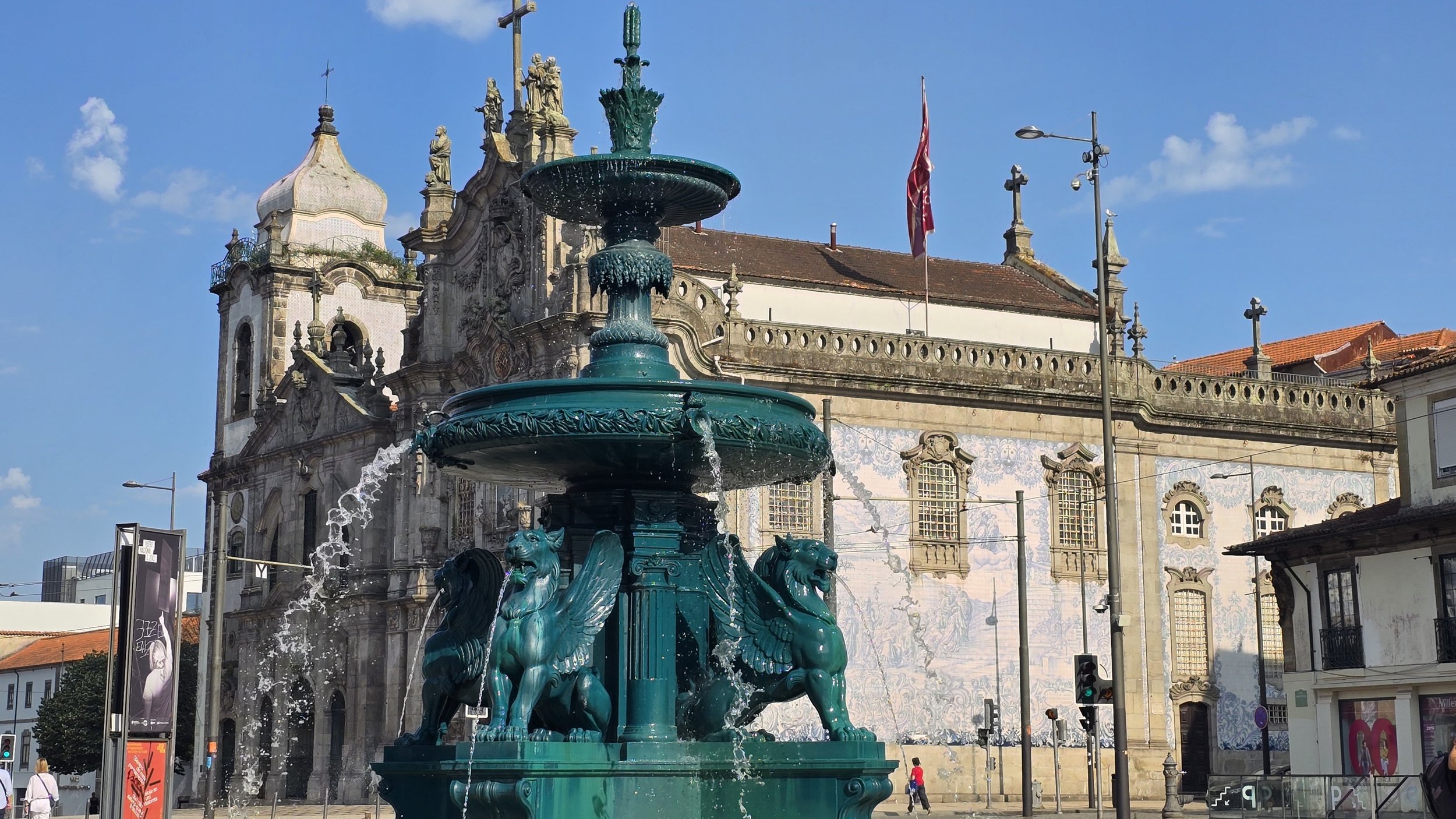 A historic building with ornate stonework and statues, a green fountain with dragon and lion sculptures in the foreground, blue sky, a flag, and streetlights.