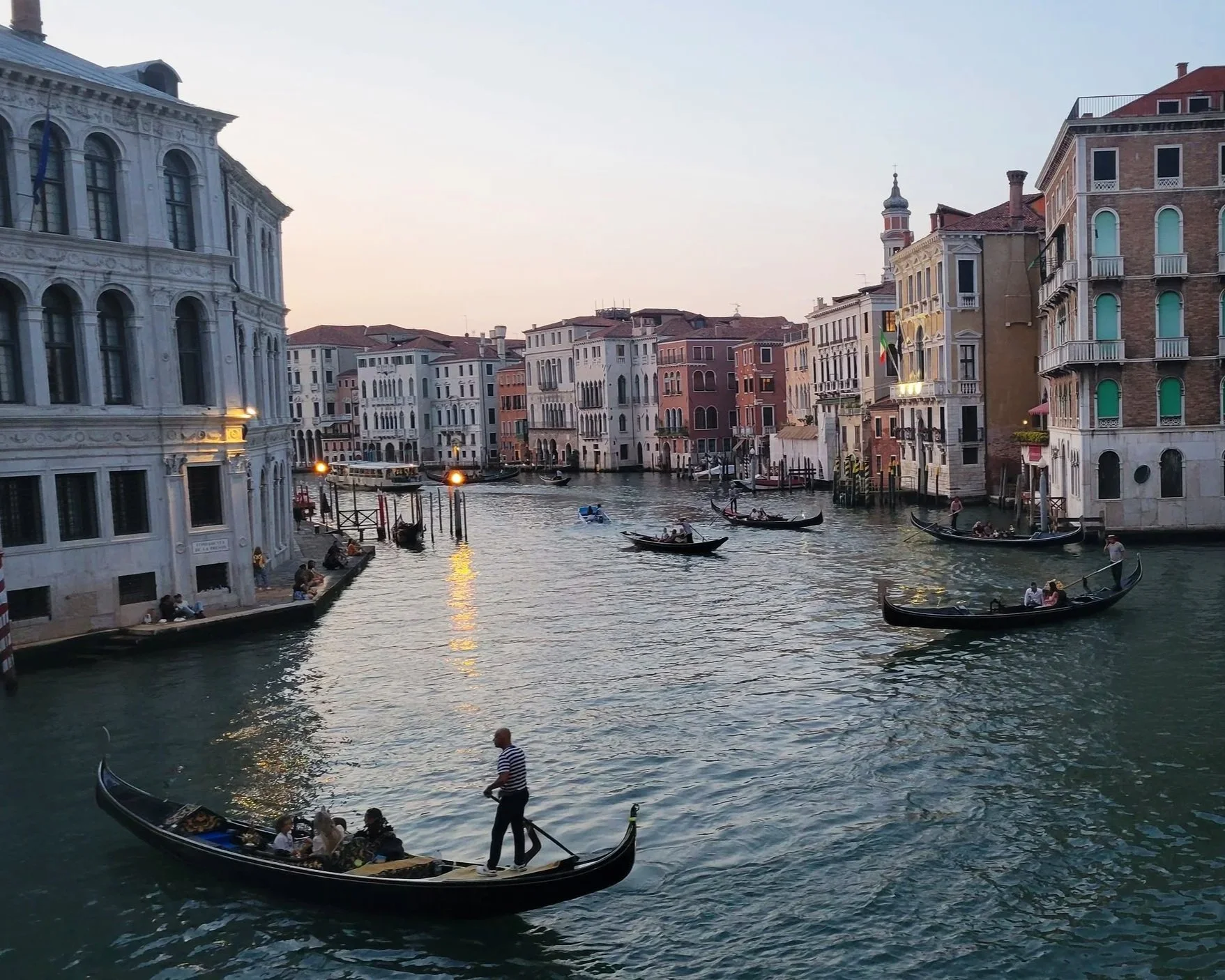 View of a canal in Venice, Italy, with gondolas carrying tourists, surrounded by historic buildings with many windows and colorful facades, under a clear sky at sunset.