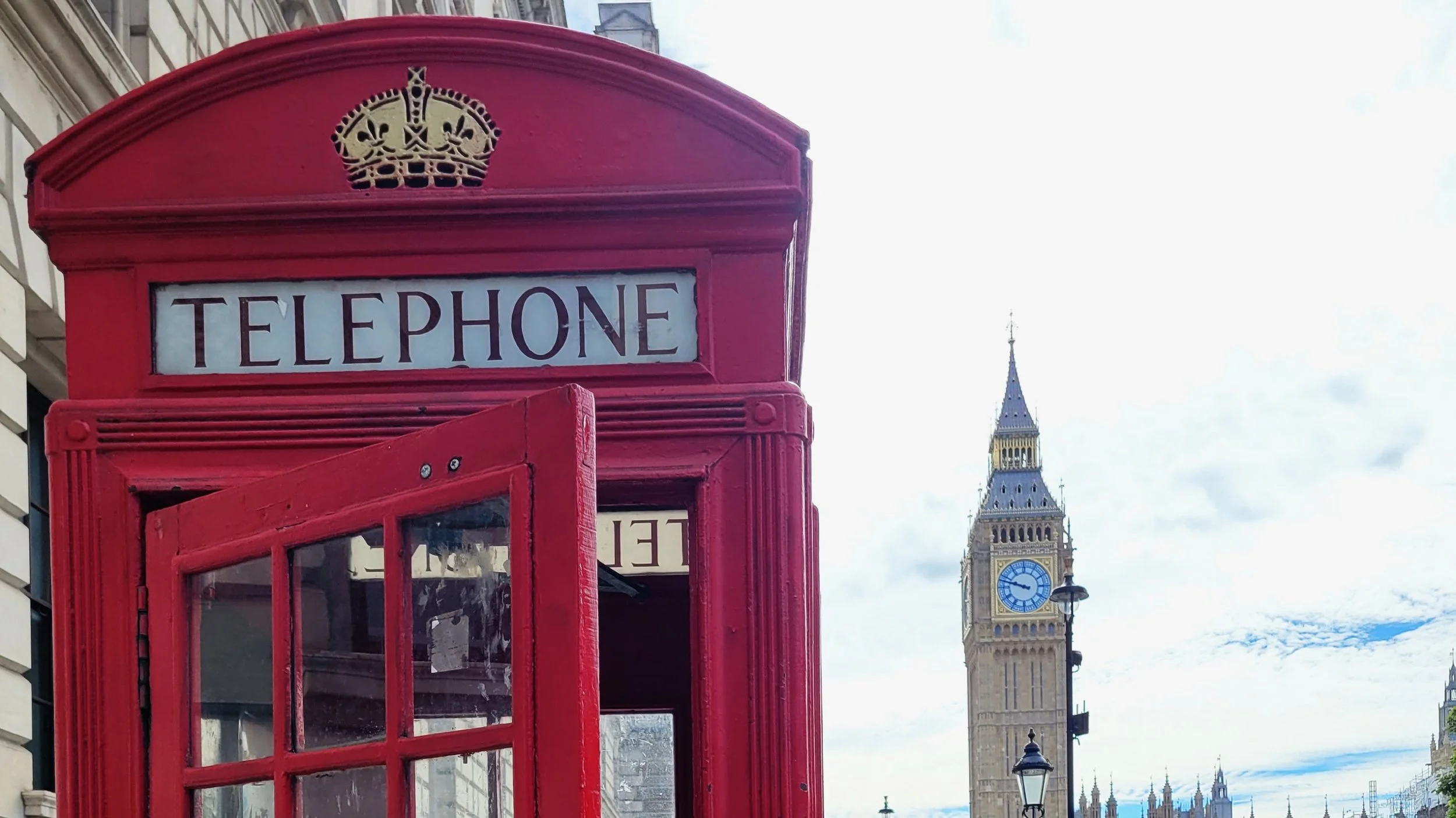 A red British telephone booth with a crown emblem on top, situated on a street near Big Ben in London, with a cloudy sky overhead.