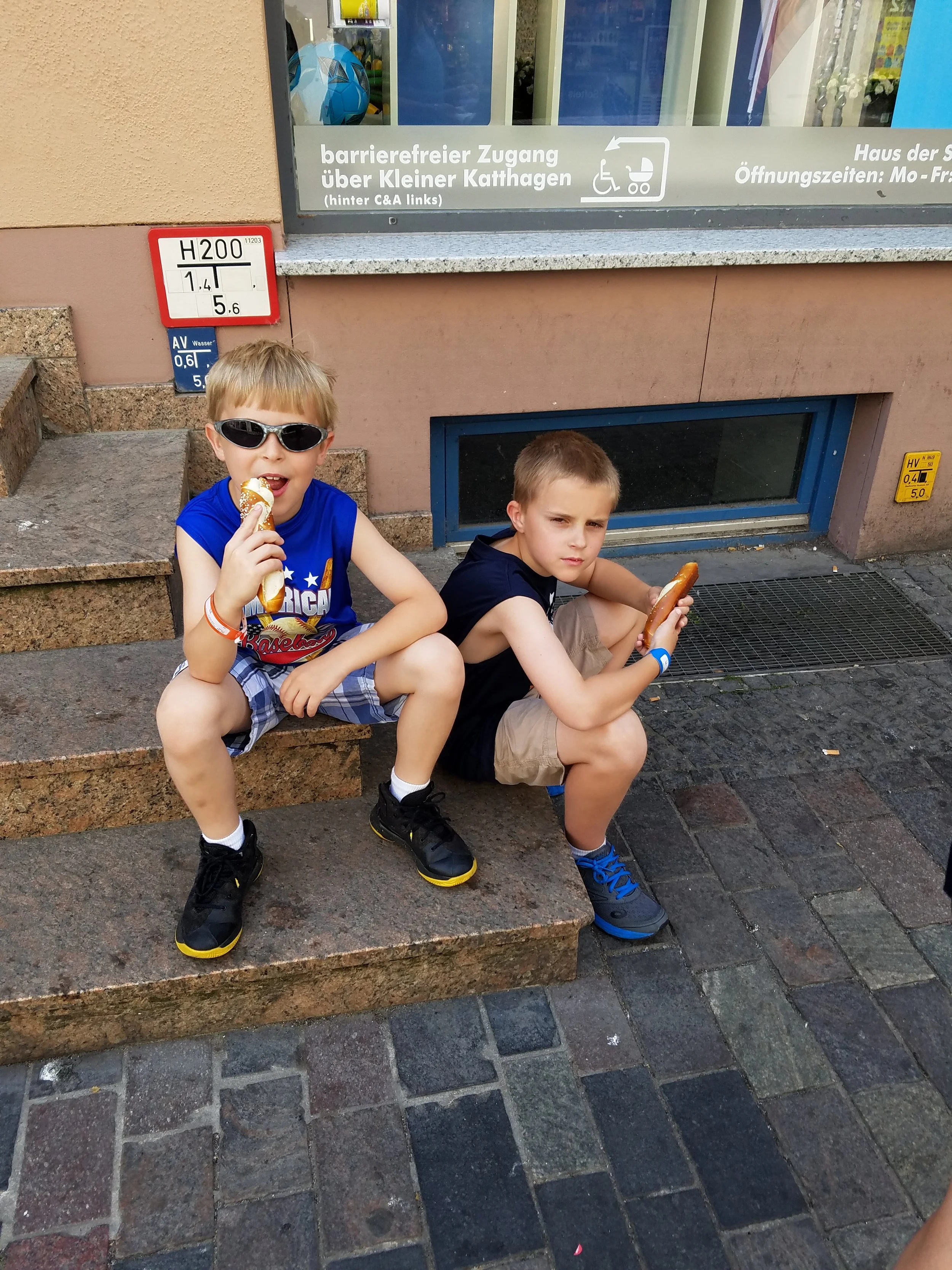 Two boys sit on stone steps along a cobblestone street in Rostock, Germany, eating large soft pretzels. A shop window and German street signage are visible on the wall behind them.