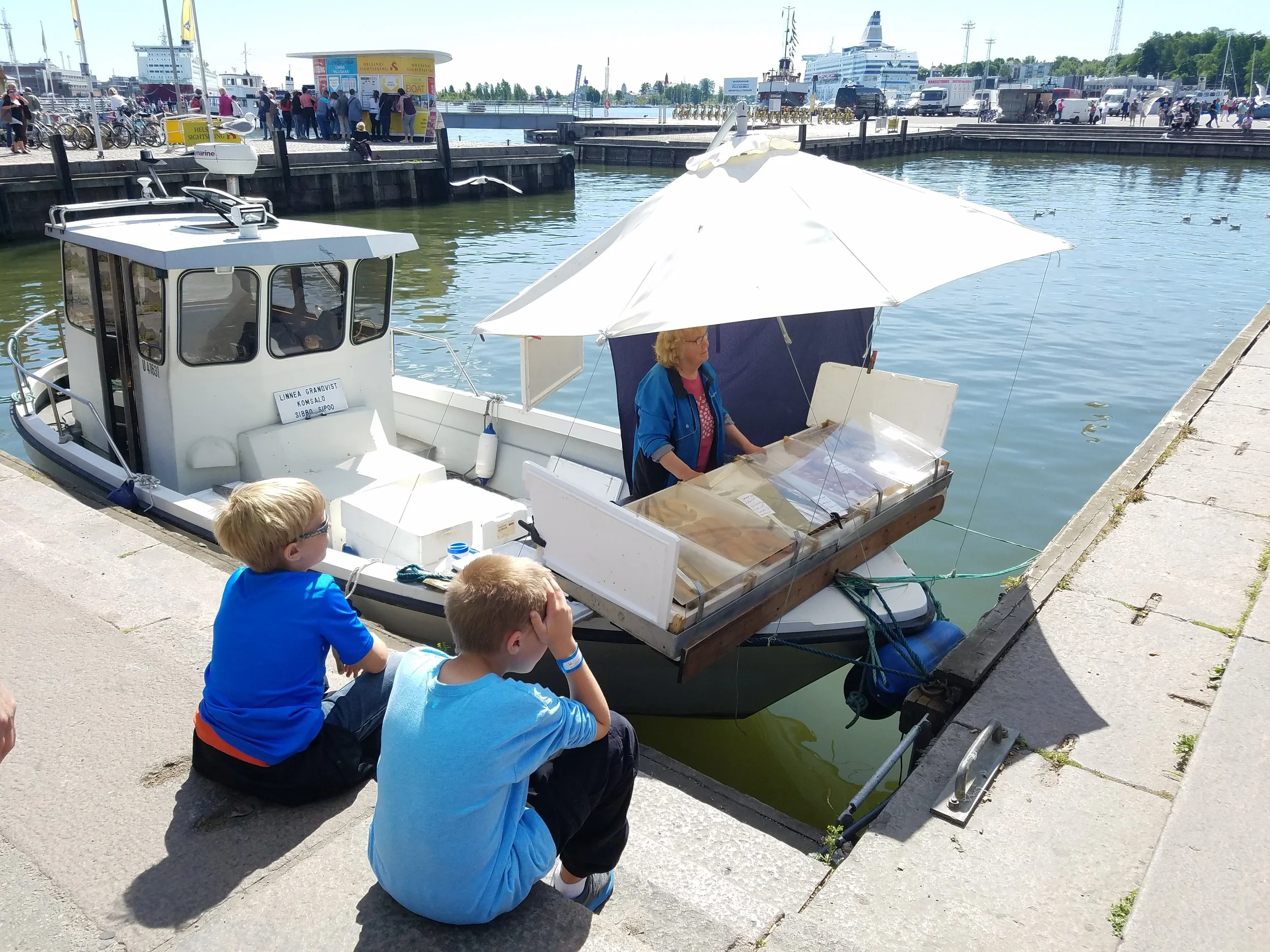 Two boys sit on a stone quay in Helsinki, Finland, watching a woman sell fish from a small boat under a white umbrella. Sightseeing boats and a ferry terminal are visible across the harbor.