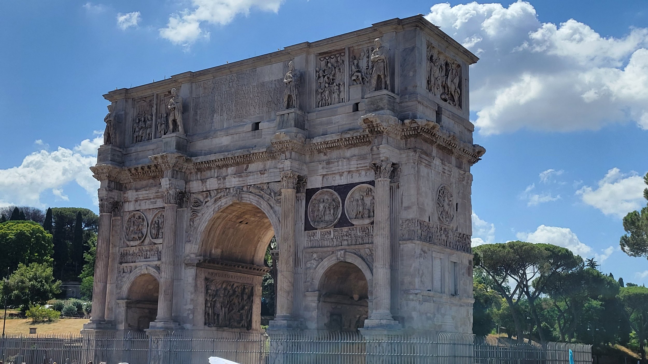 Close-up view of the Arch of Constantine in Rome, Italy, with detailed sculptures and carved reliefs, set against a blue sky with scattered clouds and surrounded by green trees.