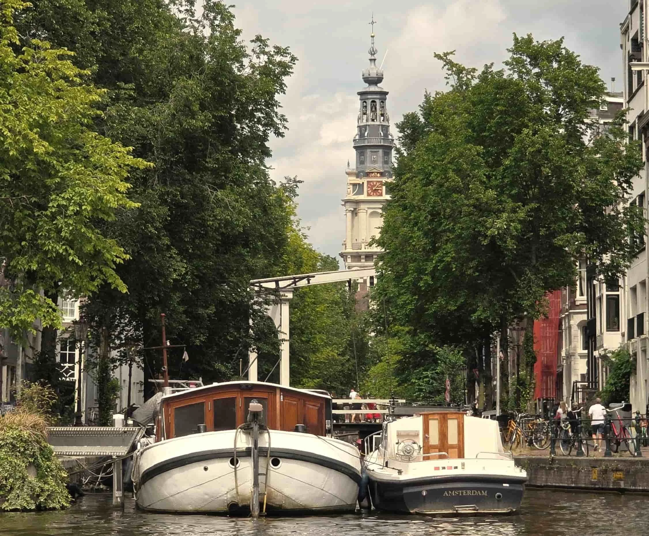 A canal with several boats docked, surrounded by lush green trees, with a historic clock tower in the background, on a cloudy day.