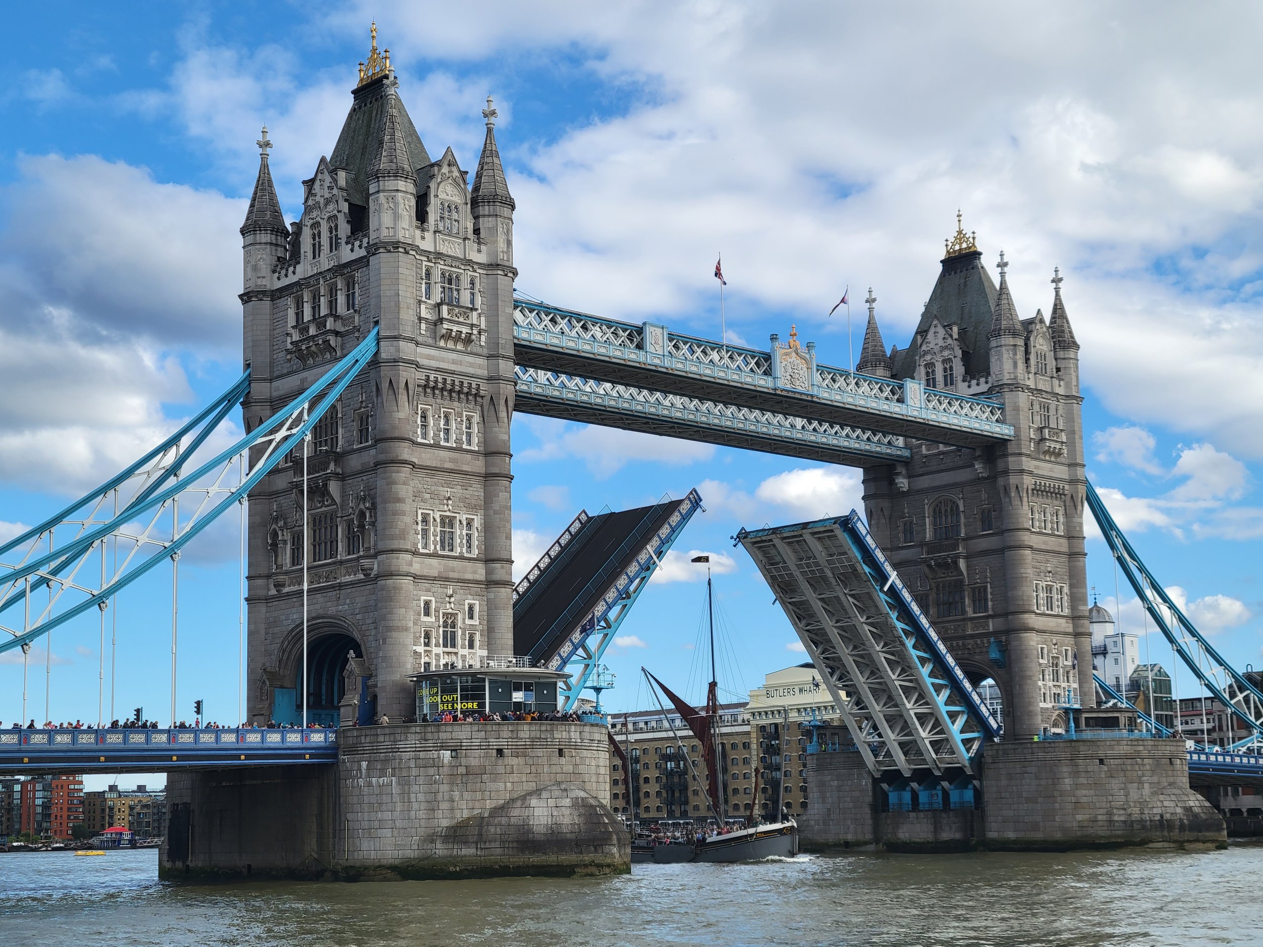 London's Tower Bridge with bascule bridges raised, boats sailing underneath, and buildings along the riverbank in the background.