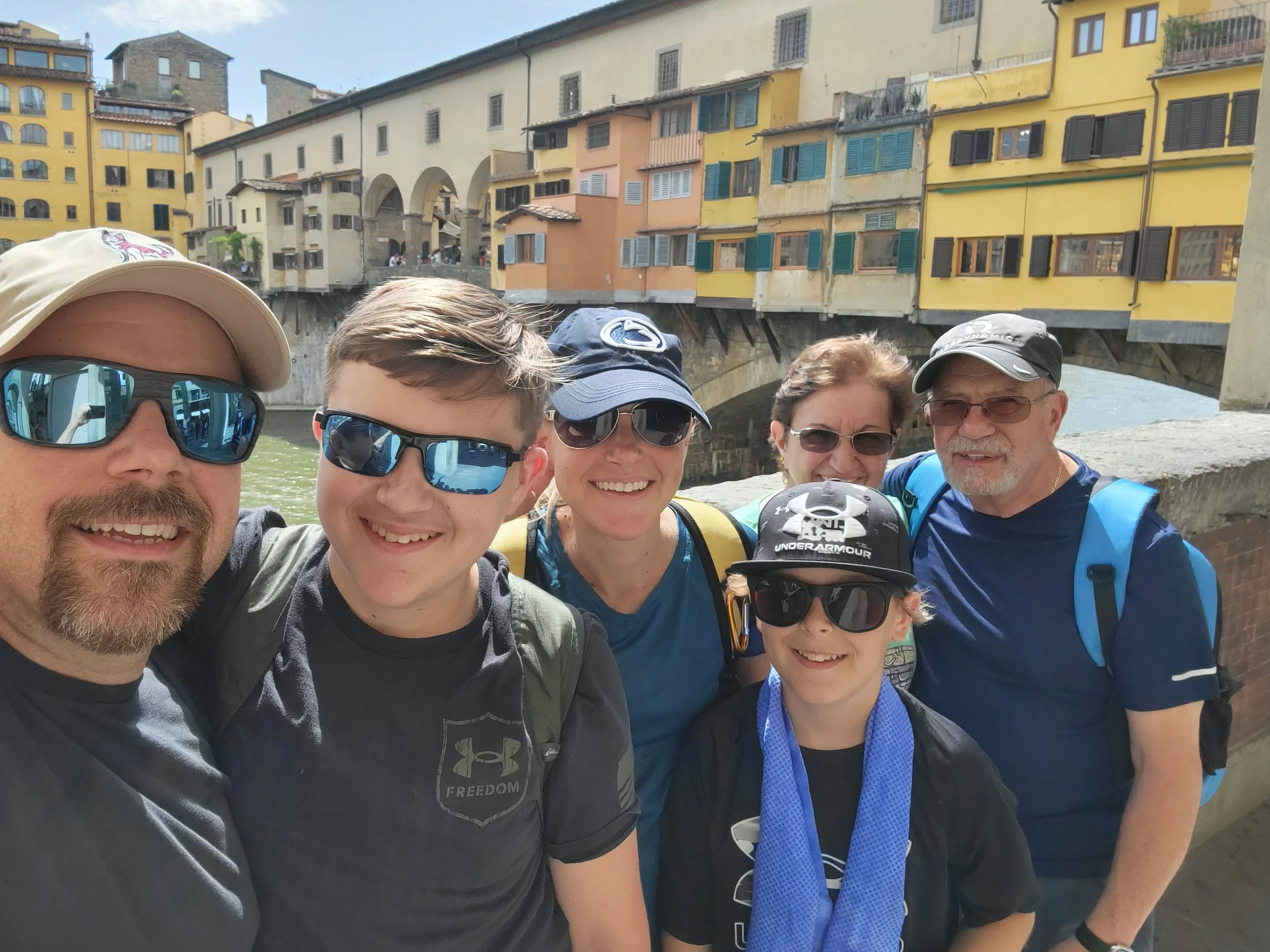 Three generations of a family taking a selfie on a bridge in Florence, Italy, with the Ponte Vecchio's colorful buildings in the background.