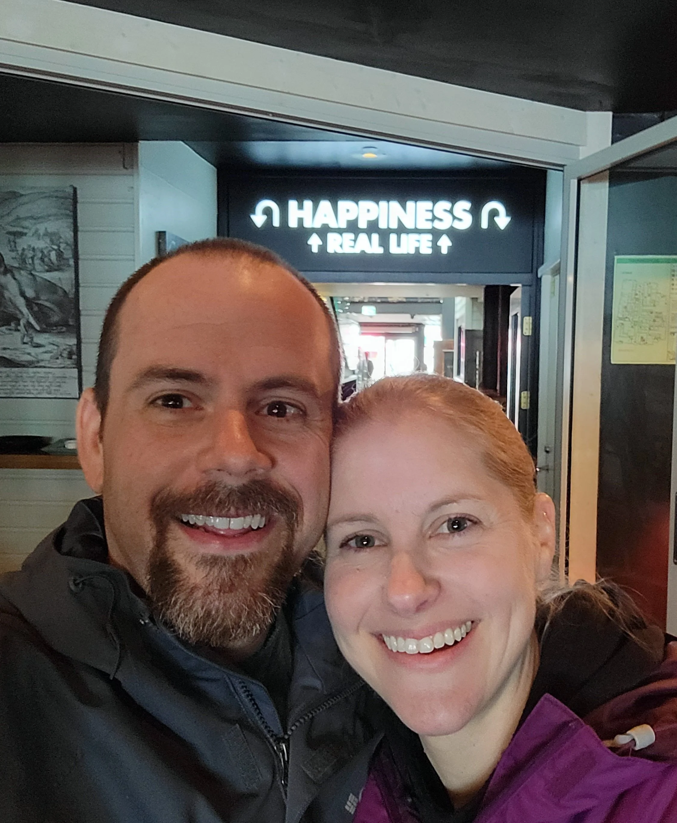 Two people smiling in a close-up photograph, with a sign in the background that reads 'Happiness Up Real Life Up' with arrows.