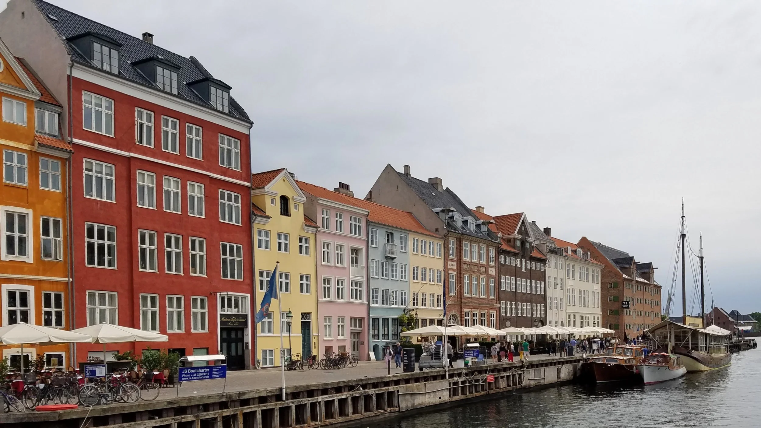 Colorful buildings along a river with boats, umbrellas, and people walking on the promenade, under a cloudy sky.