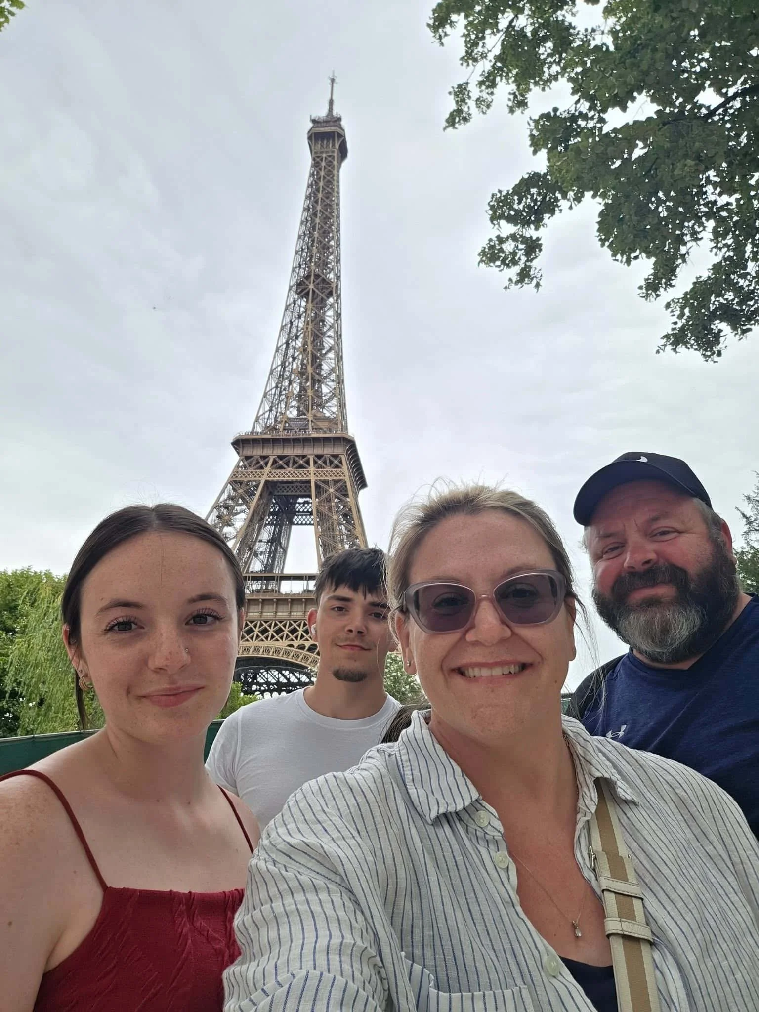 A family of four people taking a selfie with the Eiffel Tower in the background on a cloudy day. The group includes two women, one man, and a teenage boy.