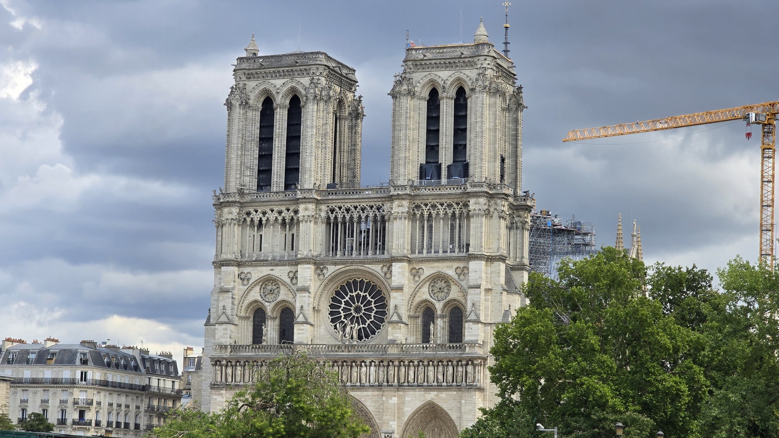 The Paris Notre-Dame Cathedral under restoration with a construction crane nearby, cloudy sky, and surrounding trees and buildings.