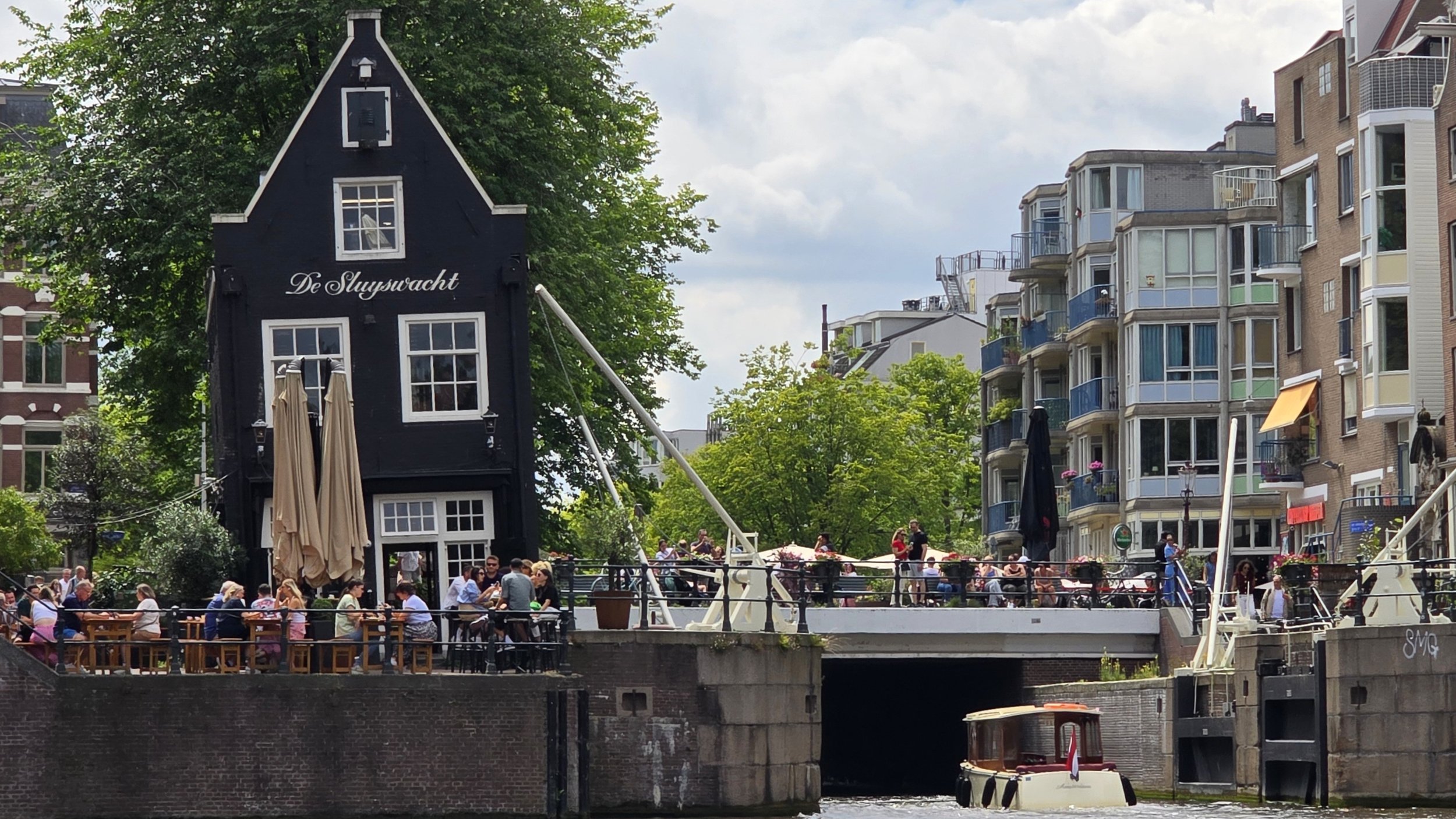 People sitting and gathering outdoors by a canal with boats, near a dark blue building labeled 'De Sluytsacht', in Amsterdam, the Netherlands with modern apartment buildings and trees.