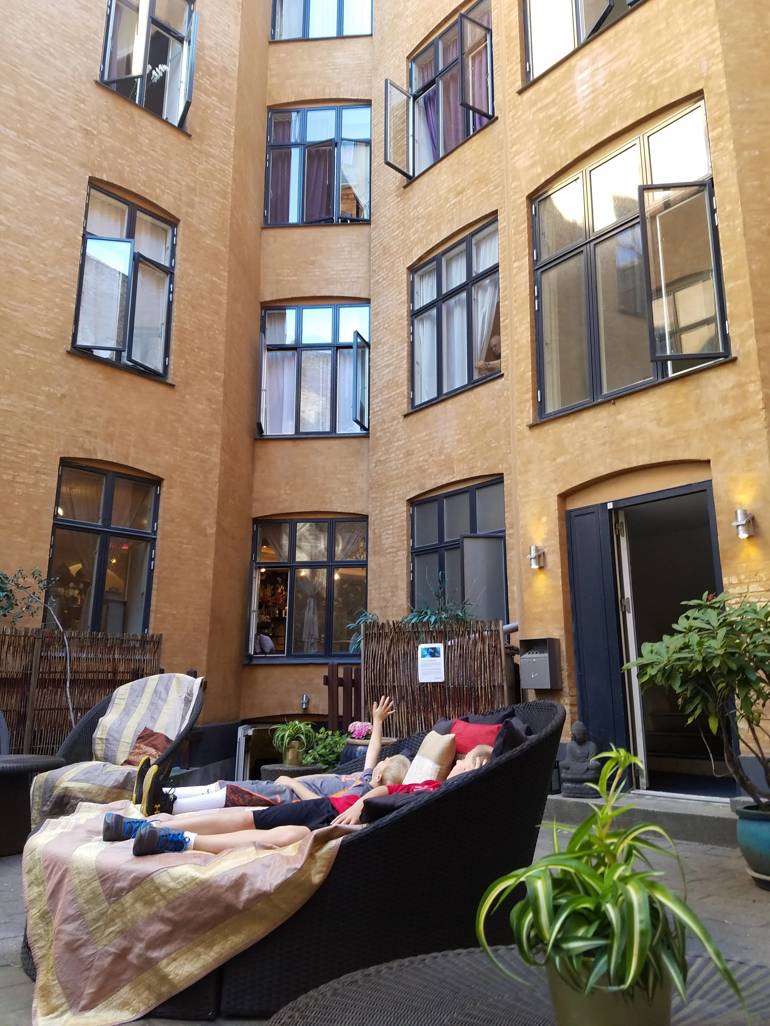 Children relaxing and playing on lounge chairs in an outdoor courtyard surrounded by a yellow brick building with large windows.