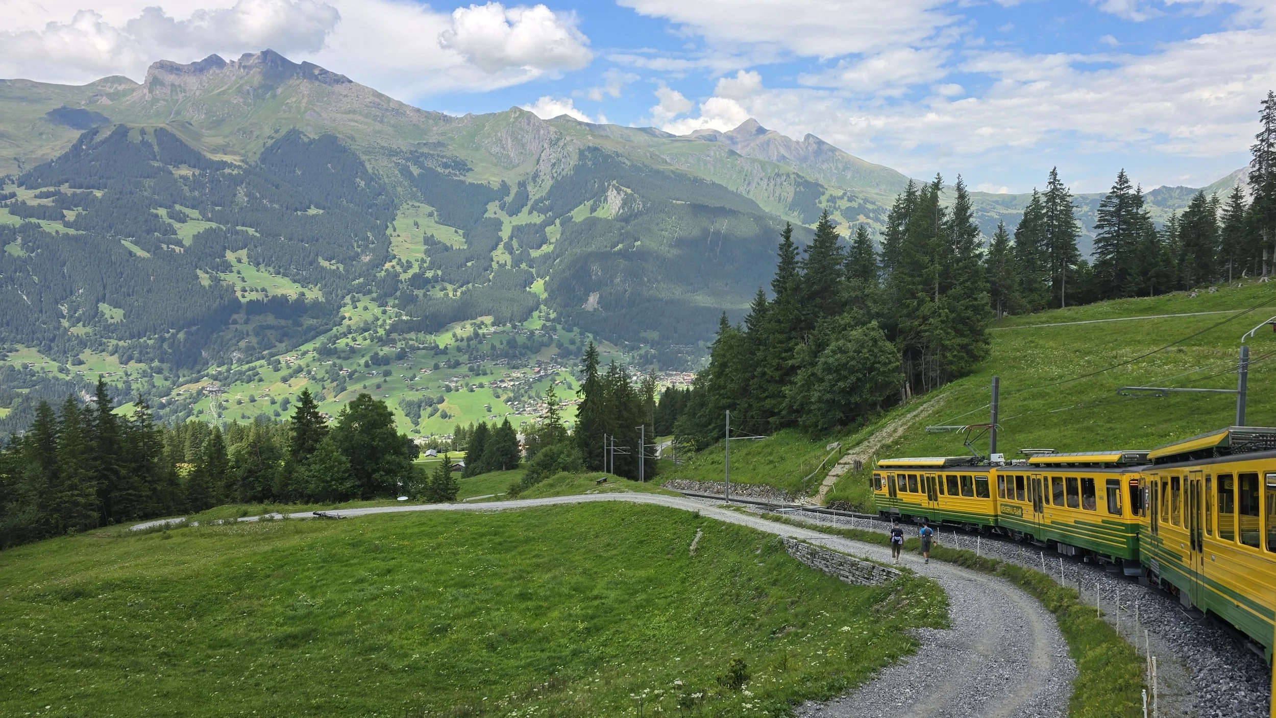 A yellow-green train traveling through a lush green mountainous landscape with trees, a village, and peaks under a partly cloudy sky.