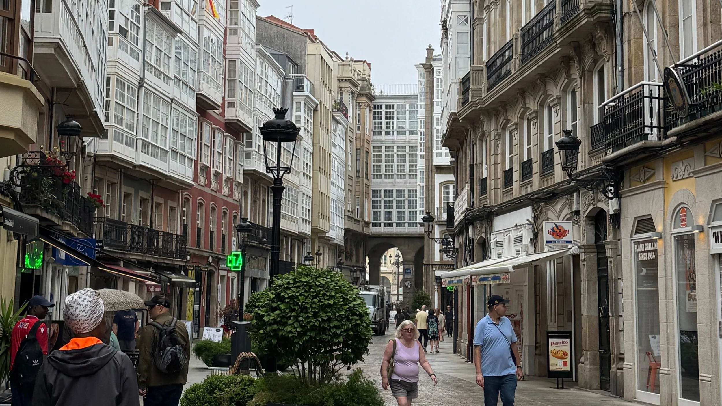 A busy city street with multiple pedestrians, decorative street lamps, and storefronts with signs. Tall buildings with ornate architecture line the street, and some people are holding umbrellas.