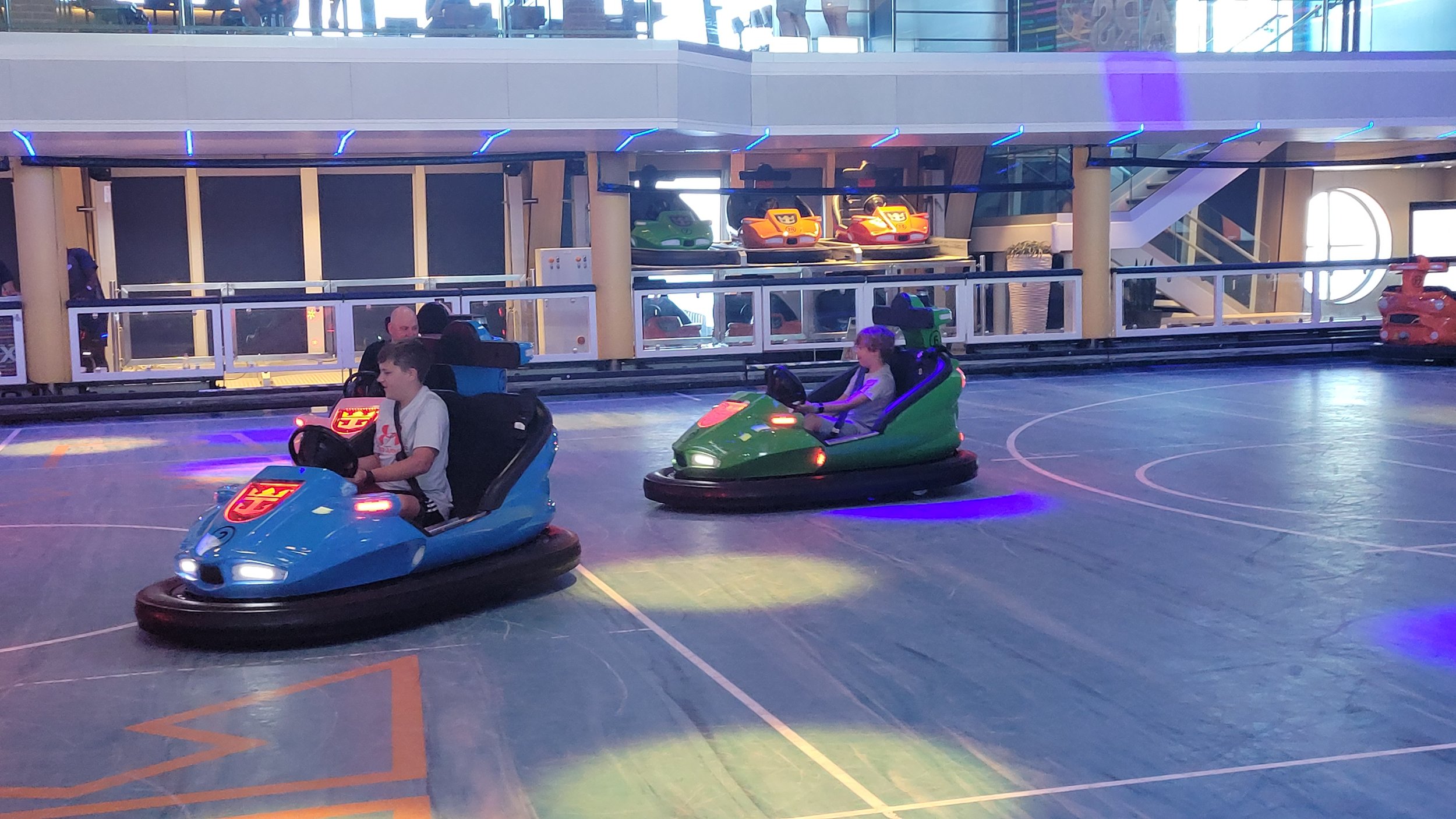 Two boys ride bumper cars on a cruise ship's indoor sports court, lit with blue and purple lights.
