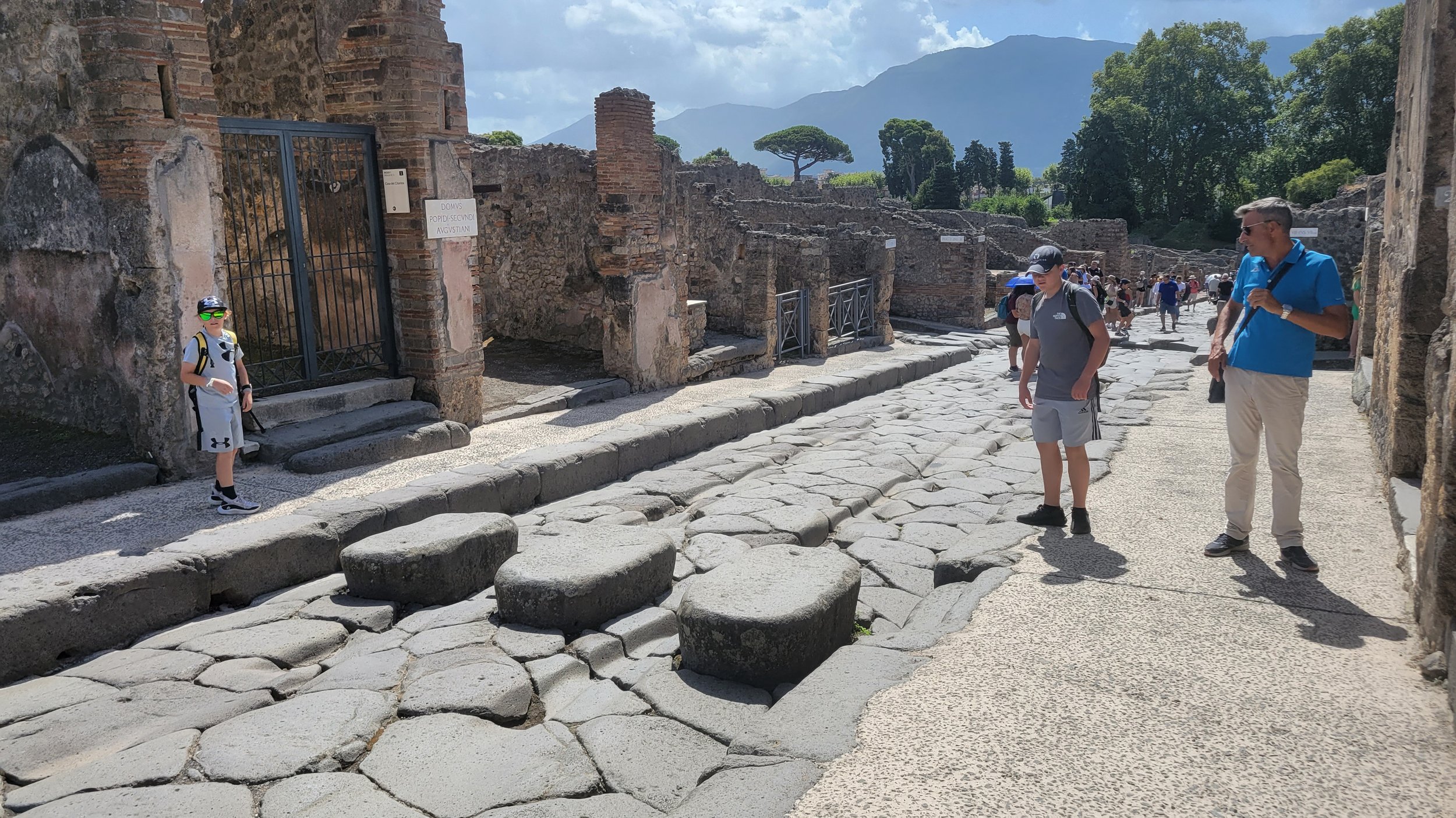 Tourists exploring ancient ruins with stone pathways, brick walls, and mountainous landscape in the background. Pompeii, Italy