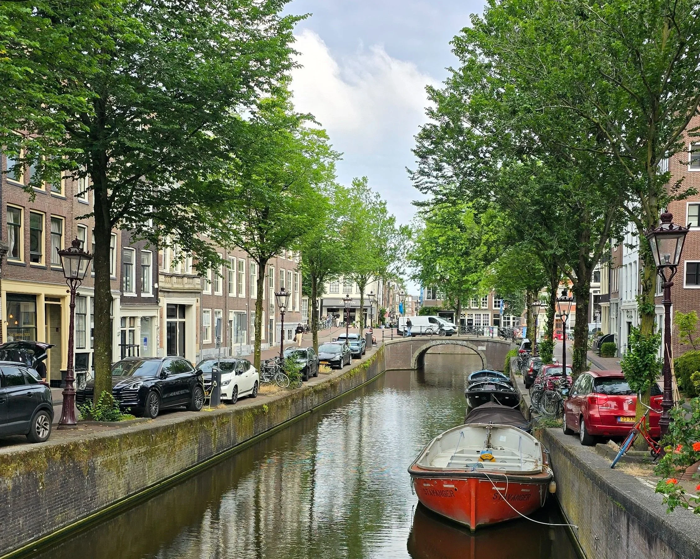 A canal lined with parked cars and bicycles, with buildings and trees on either side, and a boat floating on the water.