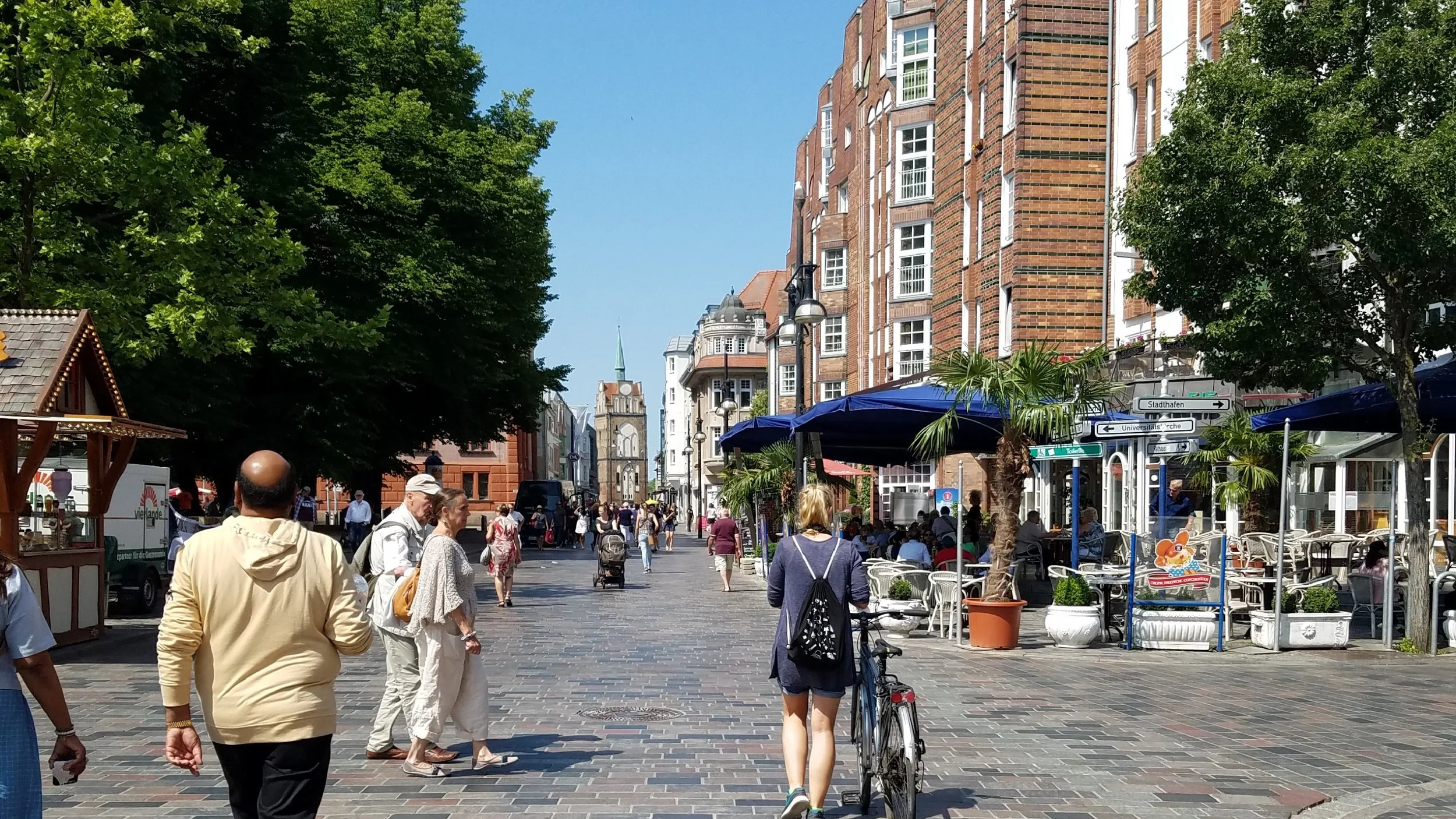 A busy city street scene with people walking and shopping. There are outdoor cafes with blue umbrellas, green trees lining the street, and historic brick buildings. A church steeple is visible in the background. Rostock, Germany