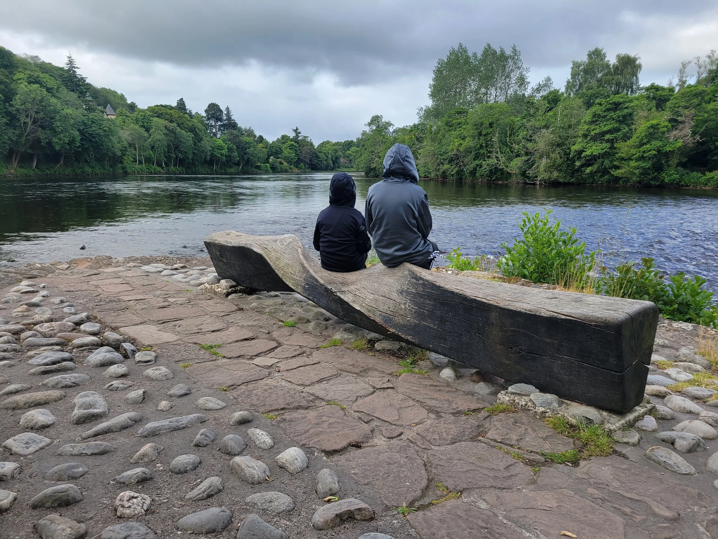 Two people sitting on a large wooden log by a river, surrounded by lush green trees, under a cloudy sky.