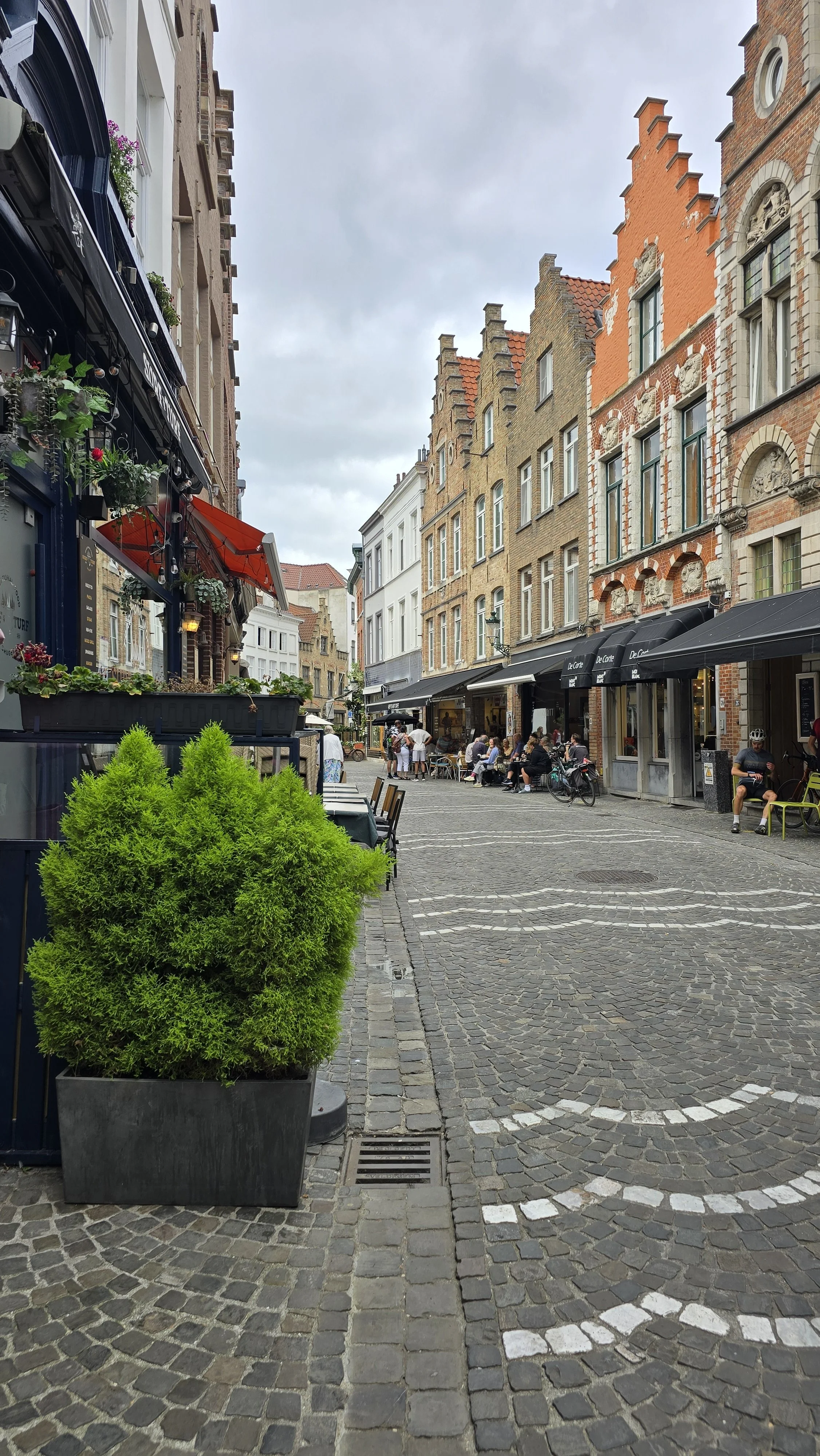 A cobblestone street in a European city with outdoor seating in front of cafes and restaurants, surrounded by historic brick and stucco buildings. There are people sitting and biking along the street, and potted plants are visible along the sidewalk.