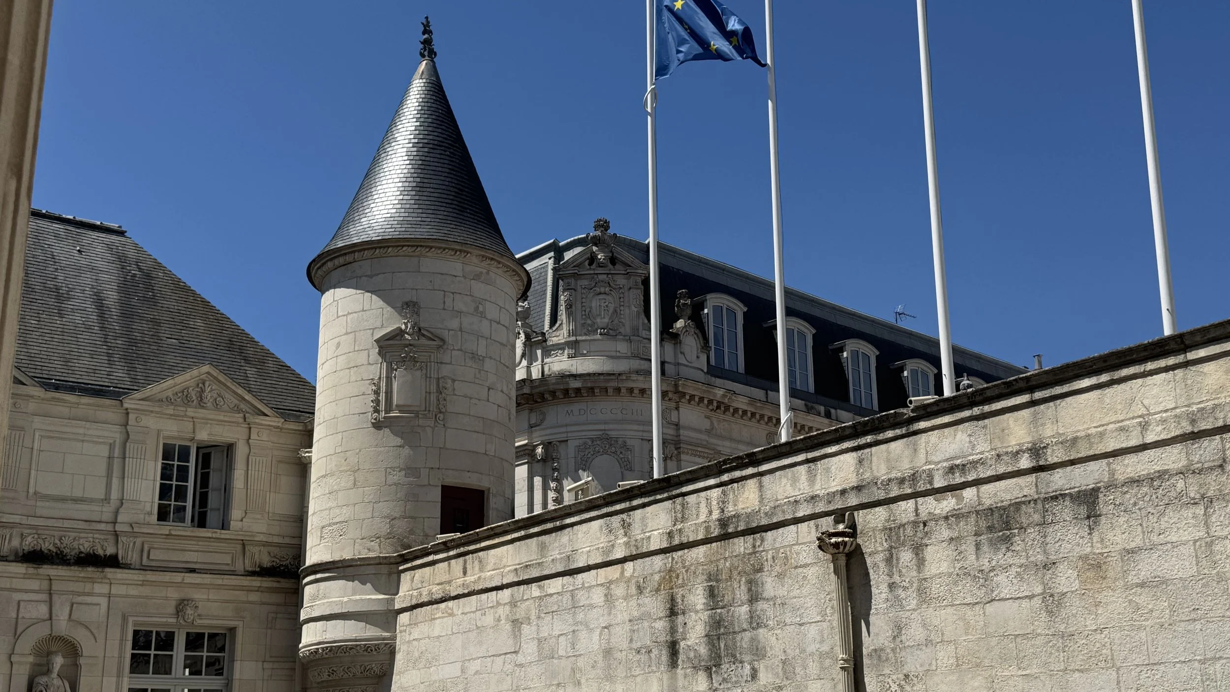 View of a historic castle or palace with detailed stone architecture, a round turret with a conical roof, and four flagpoles, one of which has an European Union flag, against a clear blue sky.