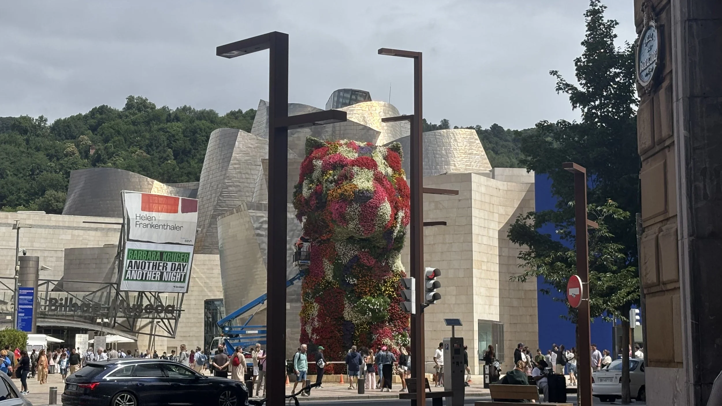 People gather outside the Guggenheim Museum in Bilbao, Spain, with a large floral sculpture in the foreground and the museum's distinctive curved architecture in the background.