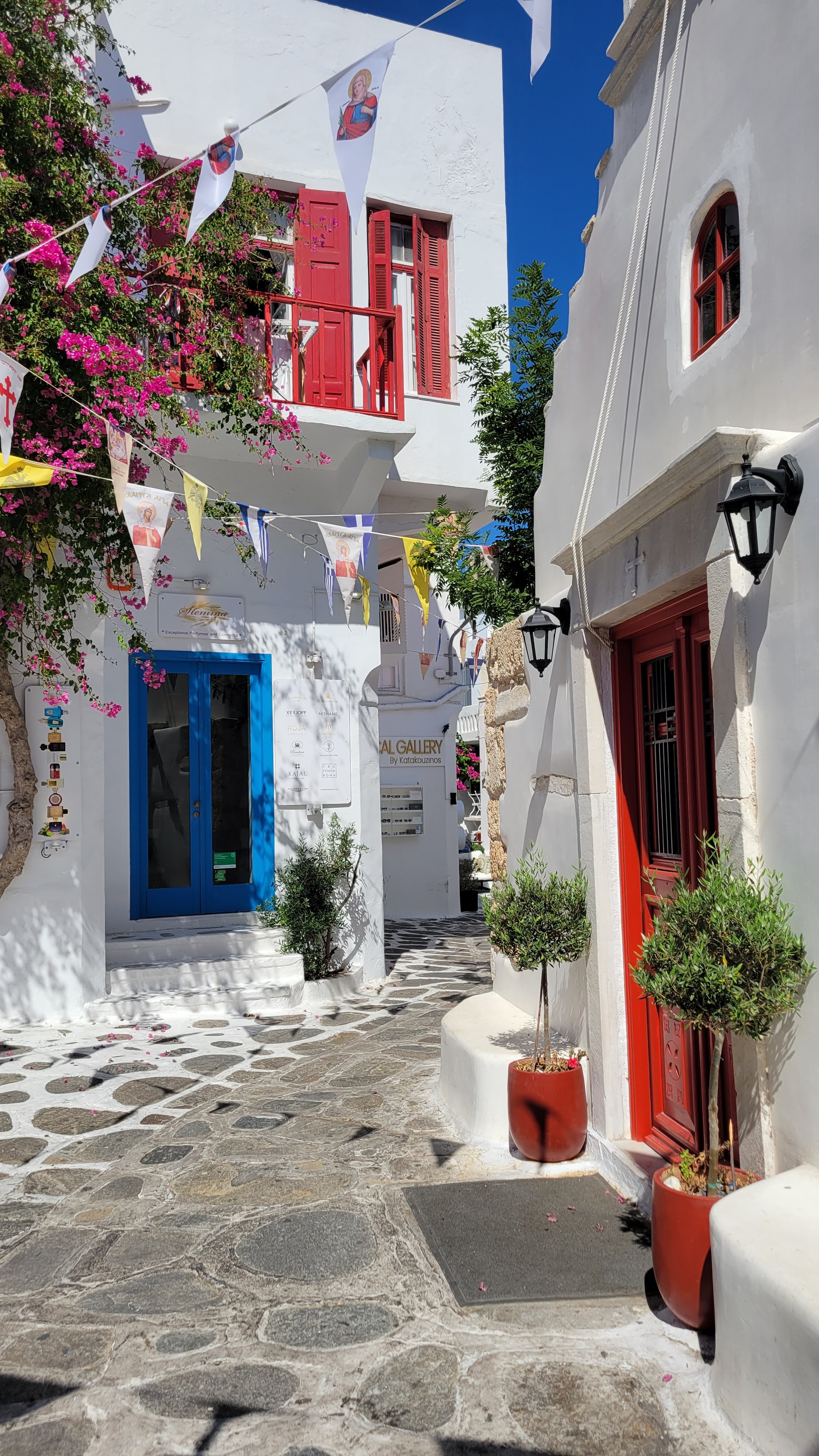 A narrow cobblestone street in a picturesque Greek village with white buildings, colorful doors, and potted plants. Decorative flags and flowers adorn the scene, with a bright blue sky overhead.