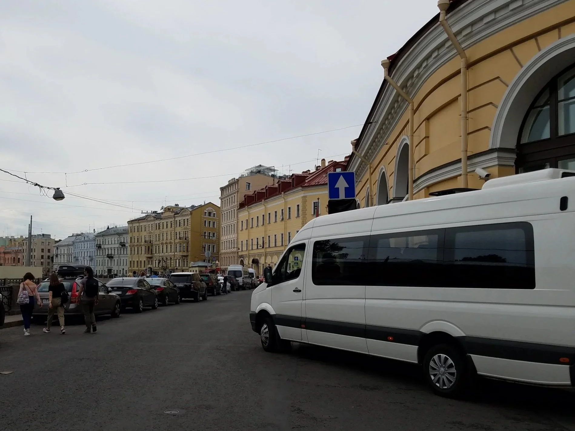 City street with historic yellow buildings, parked cars, people walking, and a white van in the foreground under cloudy sky.